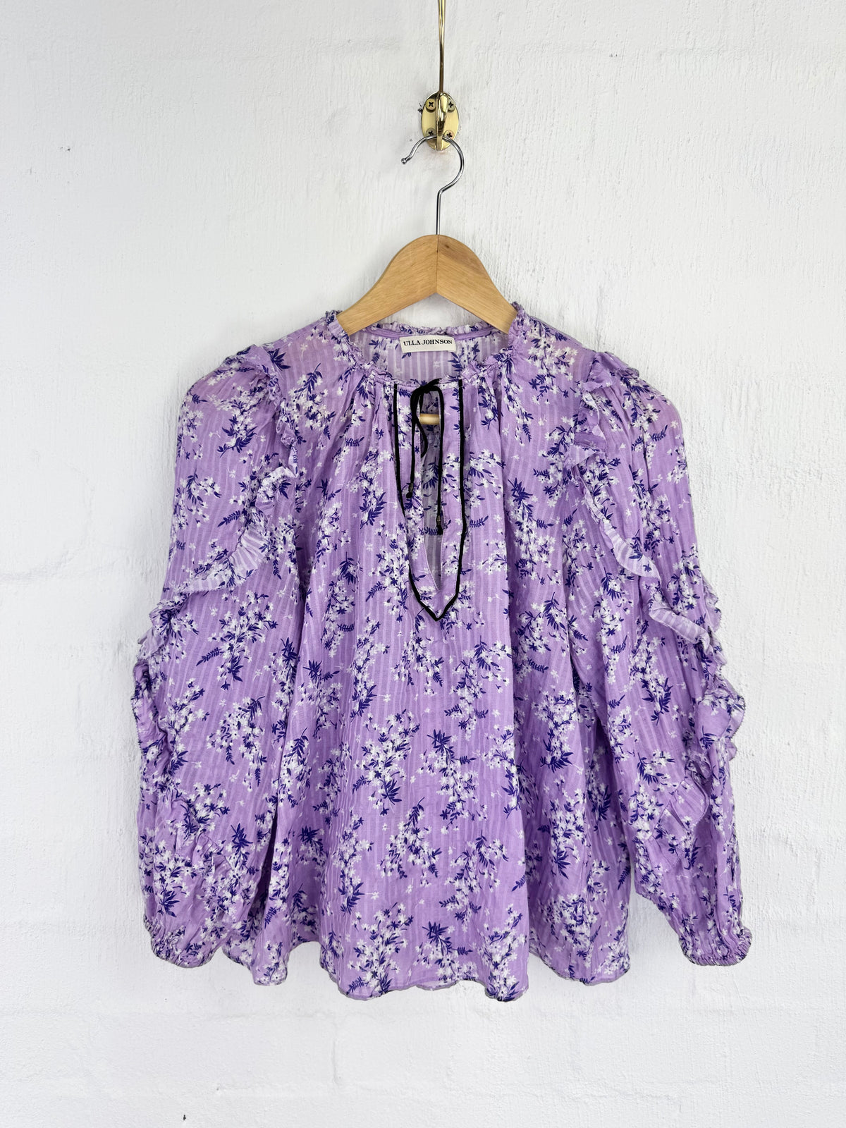 A lavender purple long-sleeve blouse with a dark purple floral print, hanging on a wooden hanger against a white painted brick wall.