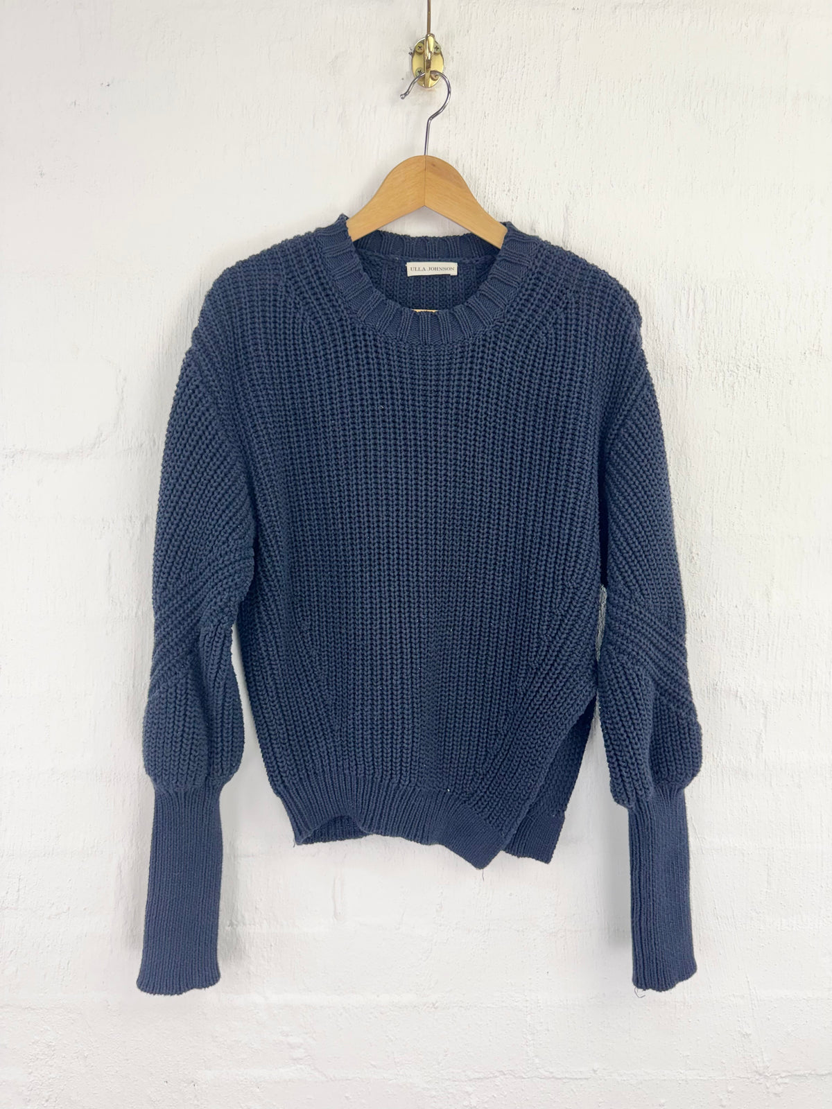 A navy blue chunky knit sweater with balloon sleeves, hanging on a wooden hanger against a white painted brick wall.