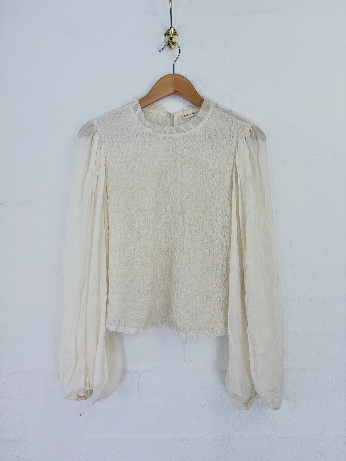 An off-white textured long-sleeve blouse with sheer balloon sleeves, hanging on a wooden hanger against a white painted brick wall.