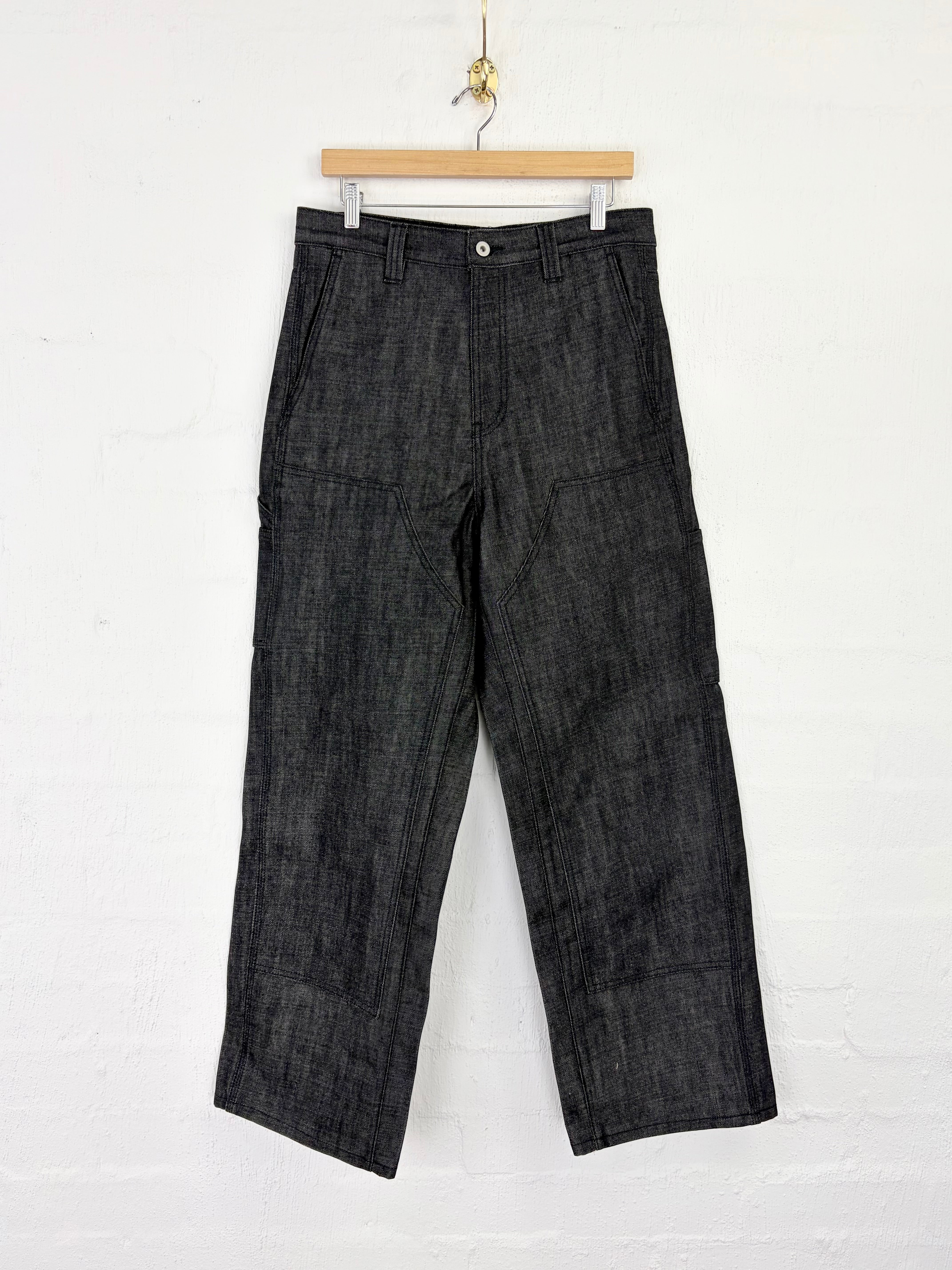 Dark charcoal gray textured pants hanging from a wooden hanger against a white brick wall.