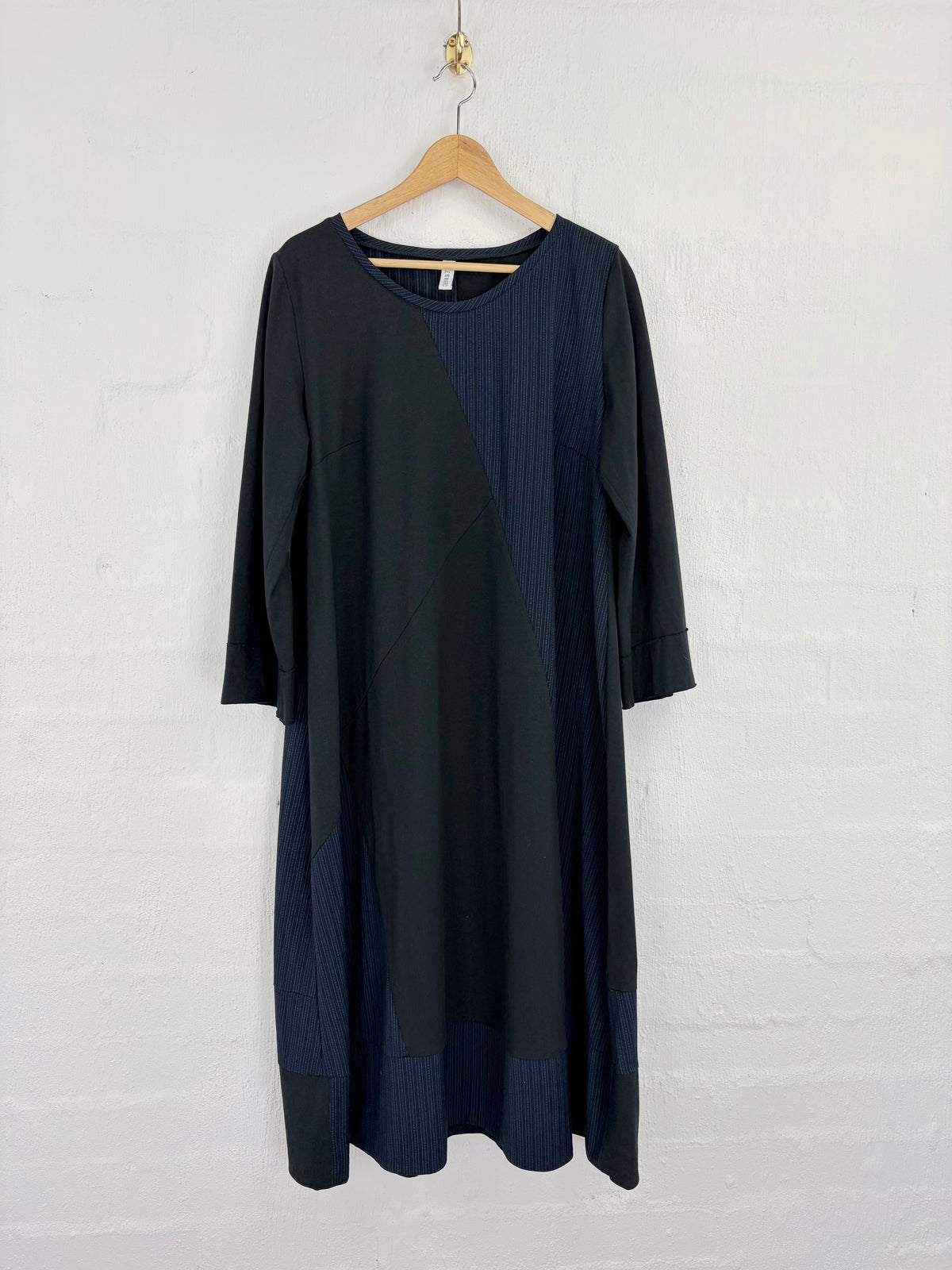 A dark navy blue dress with contrasting vertical textured panels, hanging on a wooden hanger against a white painted brick wall.