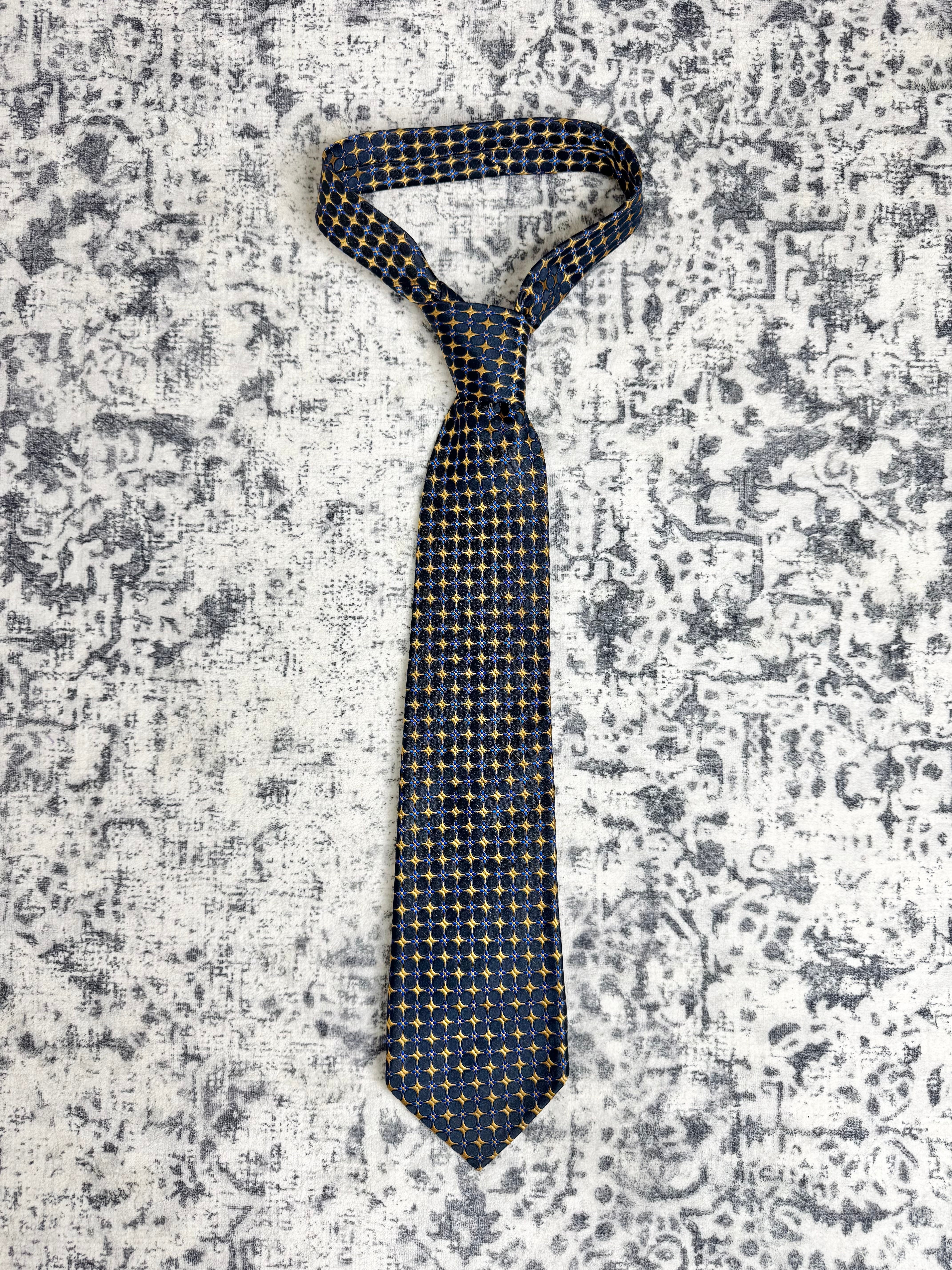 A dark navy blue necktie with a small repeating gold and white geometric pattern, laid flat on a distressed gray and white patterned background.