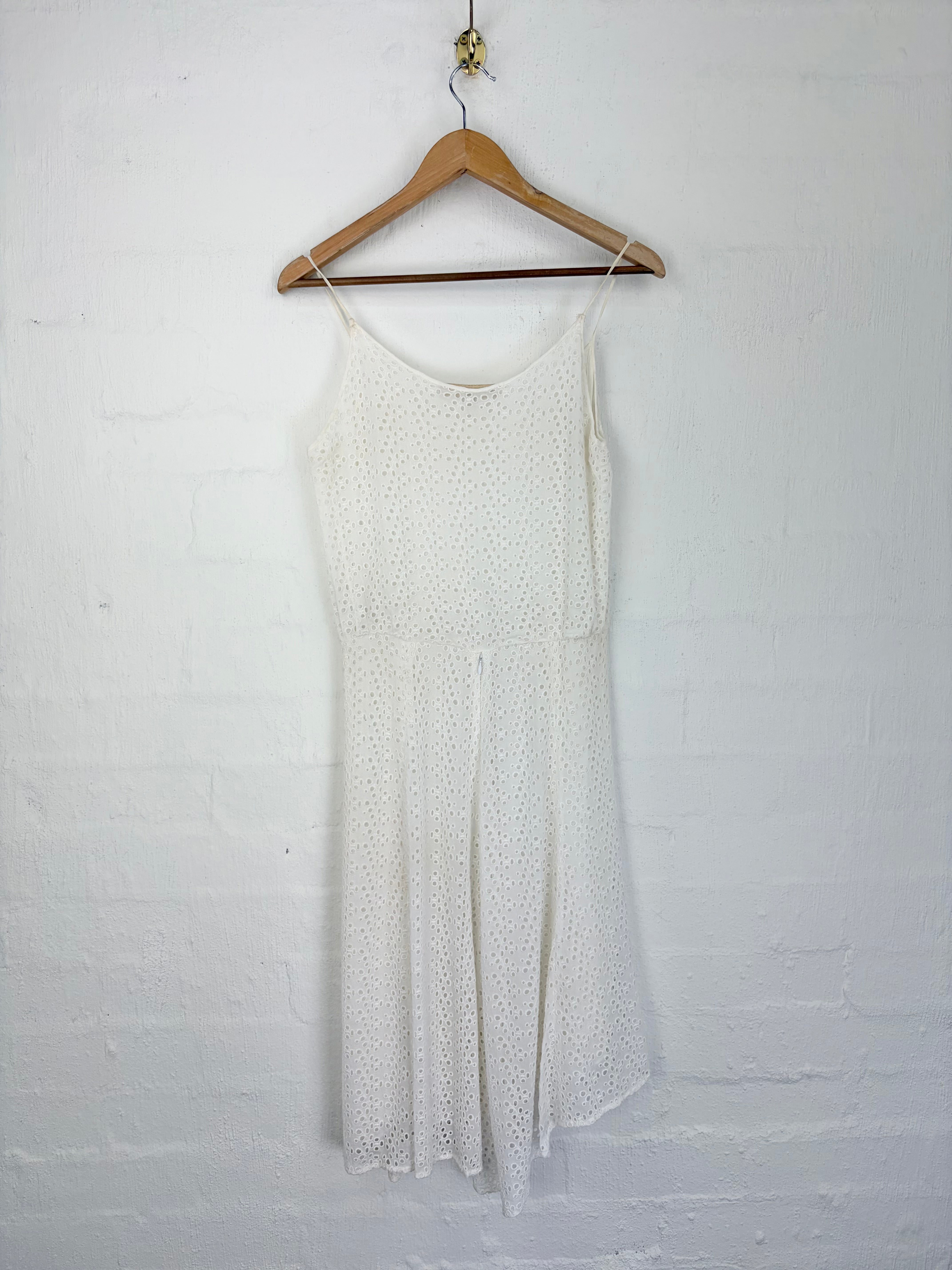 A white eyelet knit slip dress with thin spaghetti straps, hanging on a wooden hanger against a white painted brick wall.