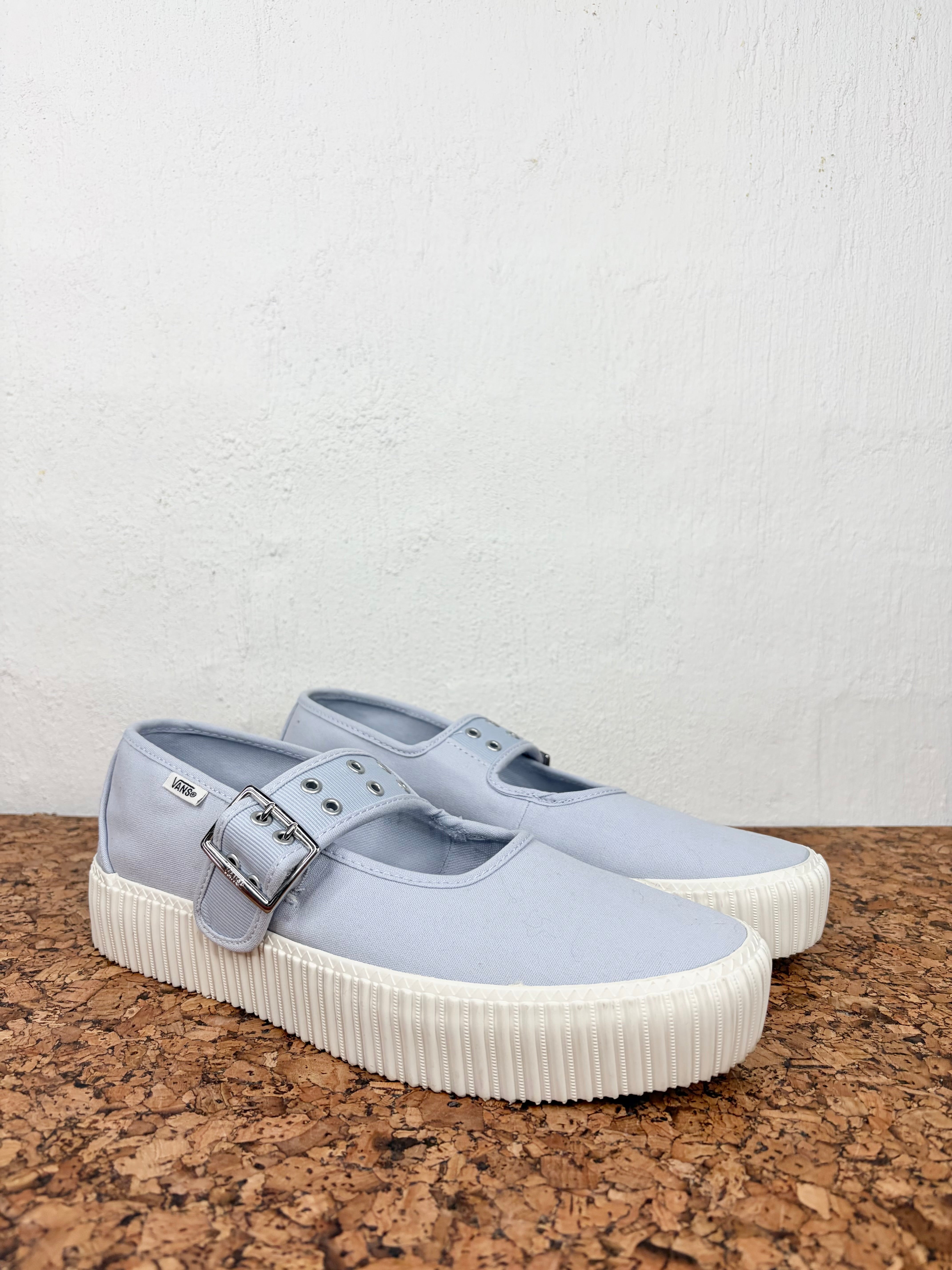 A pair of light blue canvas Mary Jane platform sneakers with silver buckle straps and thick white pleated soles, displayed on a cork surface against a white wall.