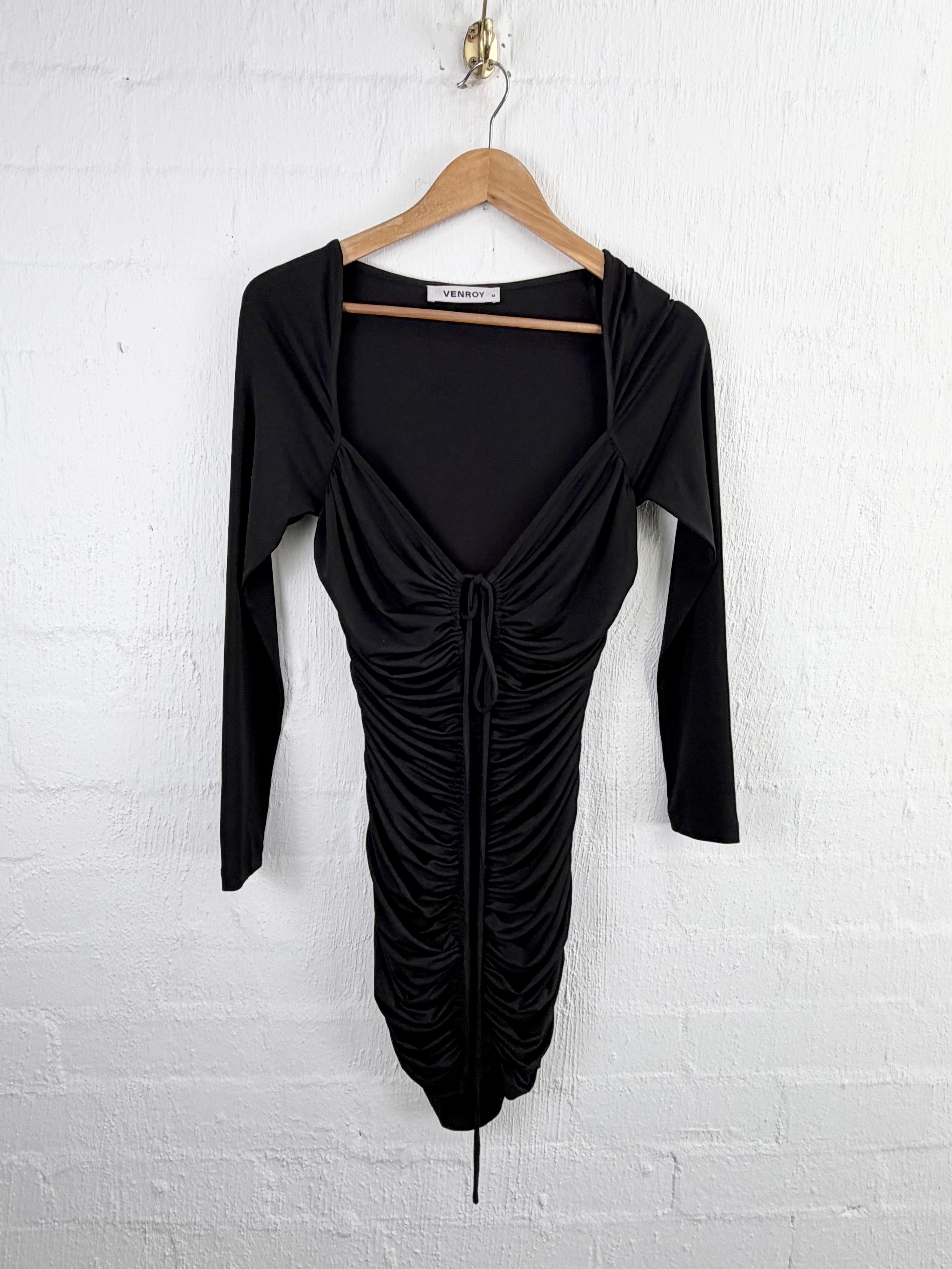 A black long-sleeve ruched dress hanging on a wooden hanger against a white brick wall.