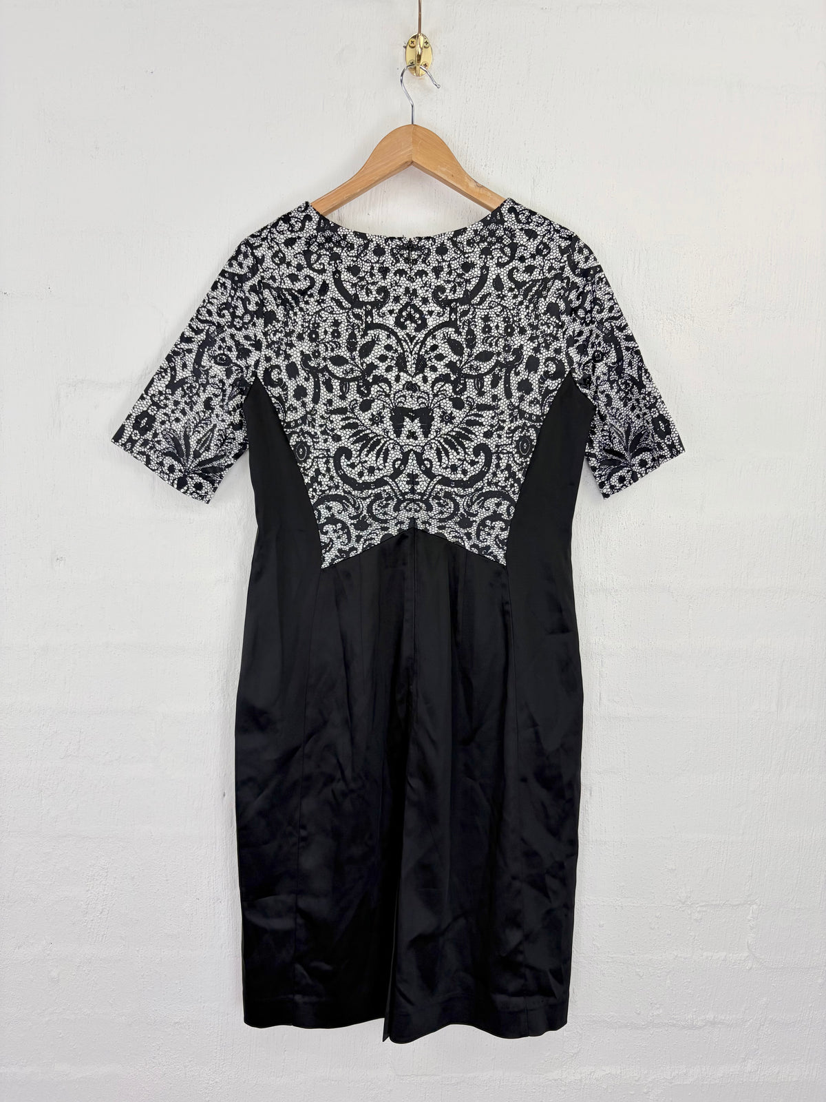A black and white patterned short-sleeve dress hanging on a wooden hanger against a white brick wall.