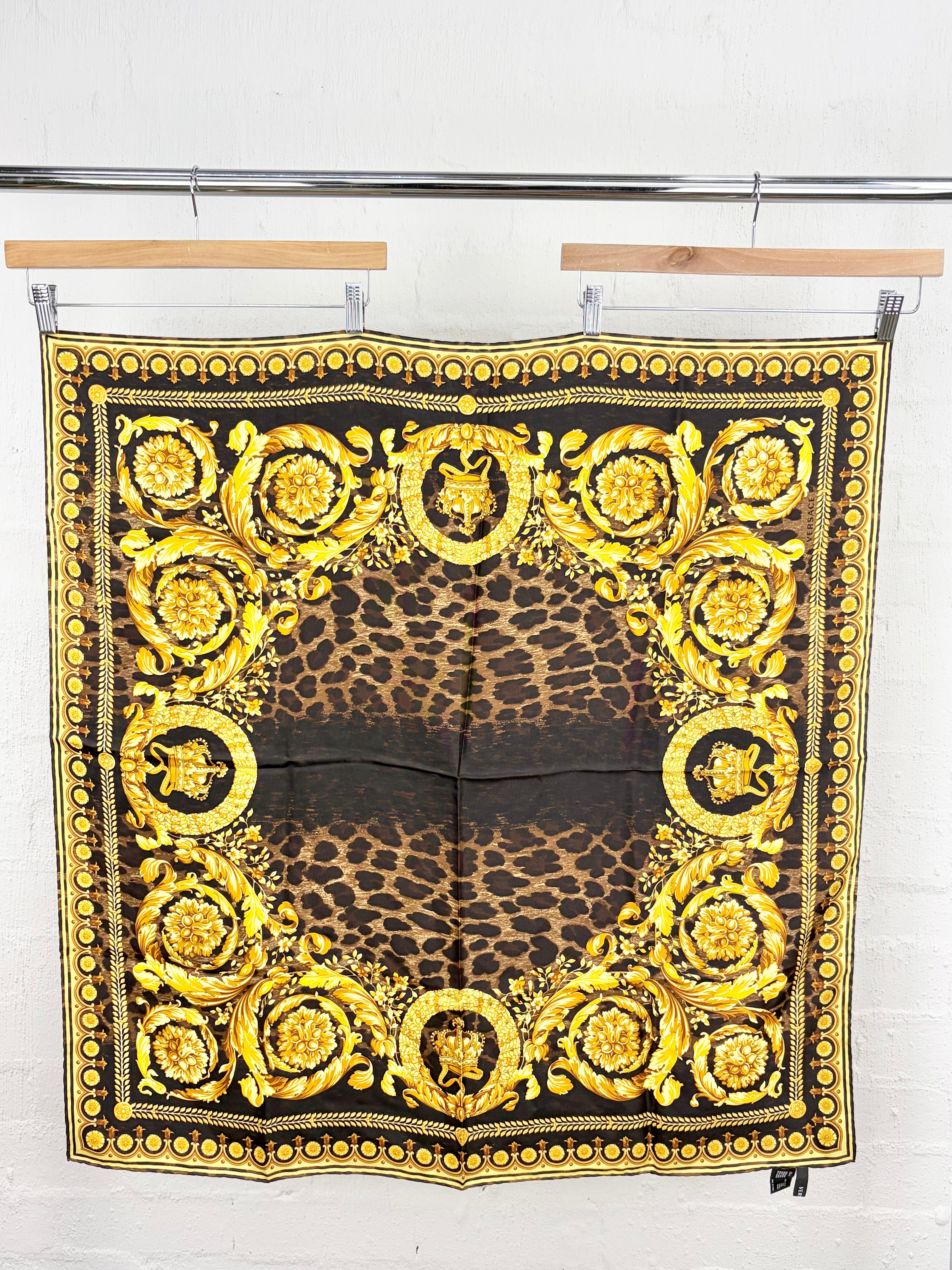 A square Versace scarf with a leopard print center and ornate gold baroque medallions and borders, hanging from wooden hangers on a metal rack against a white brick wall.