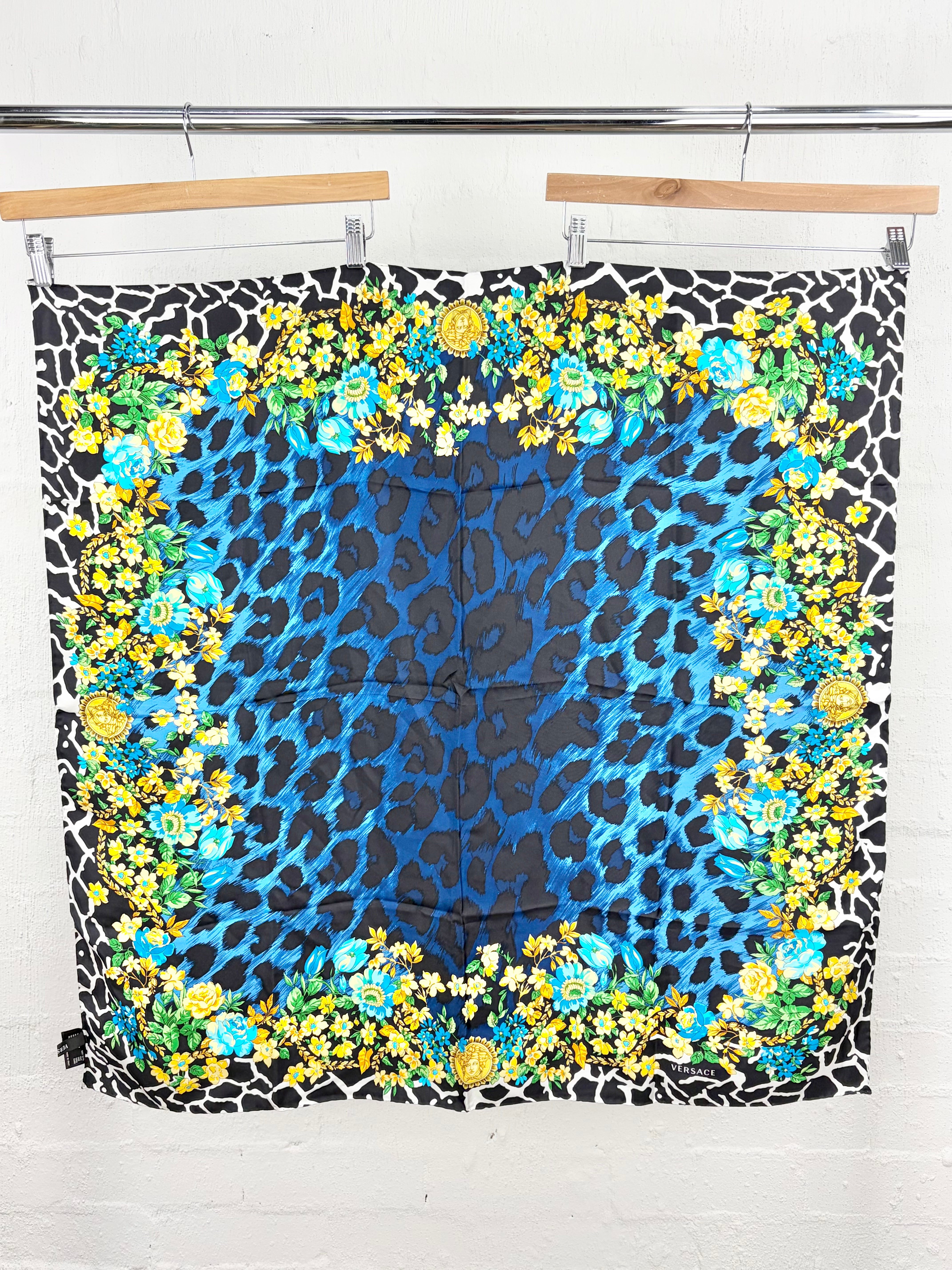 A square scarf with a vibrant blue leopard print center, surrounded by a border of yellow and blue floral motifs, hanging from wooden hangers against a white brick wall.