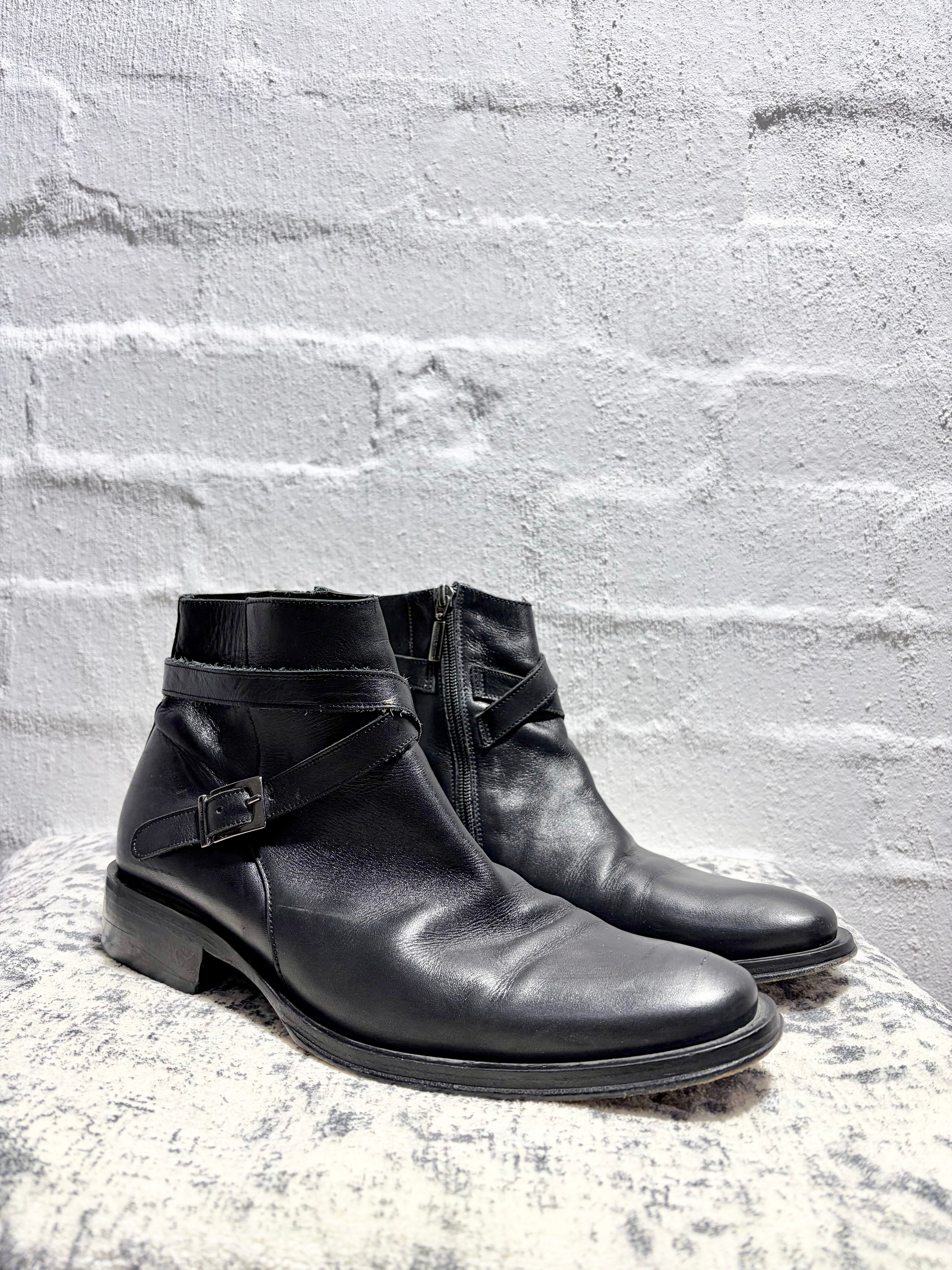 A pair of black leather ankle boots with buckle straps, displayed on a textured stone surface against a white brick wall.