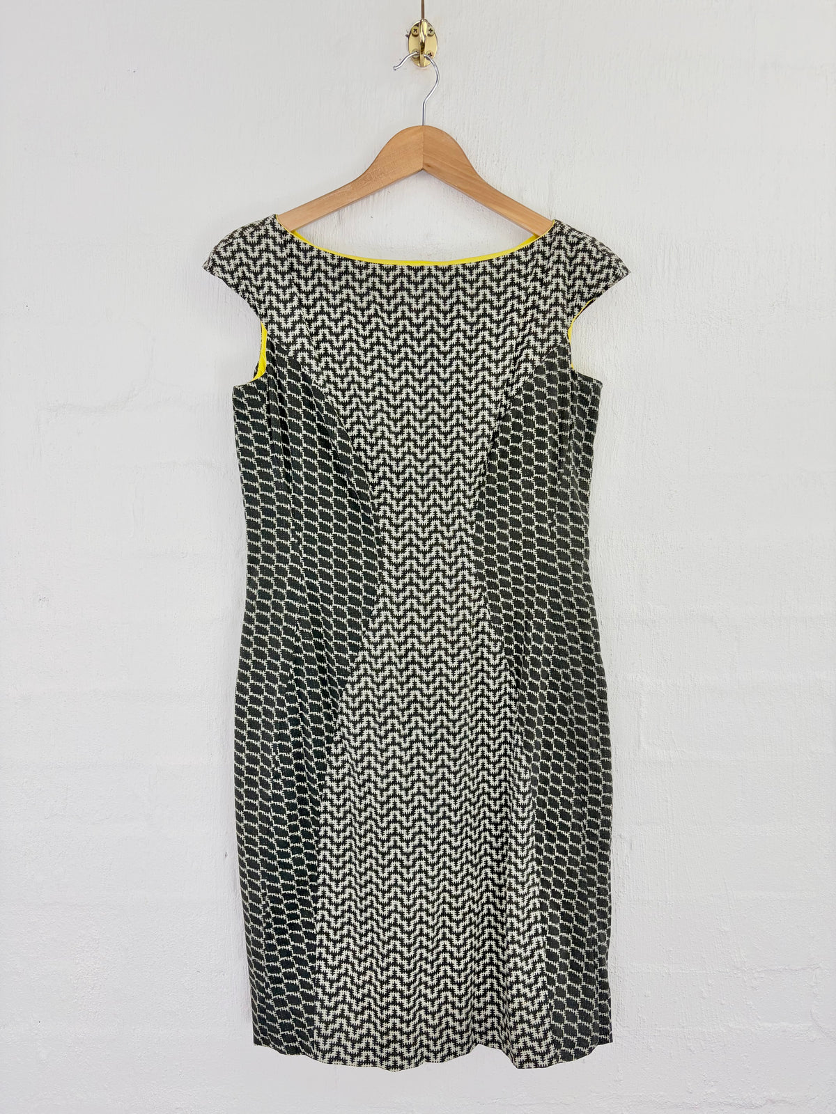 A sleeveless black and white geometric pattern dress hanging on a wooden hanger against a white brick wall.