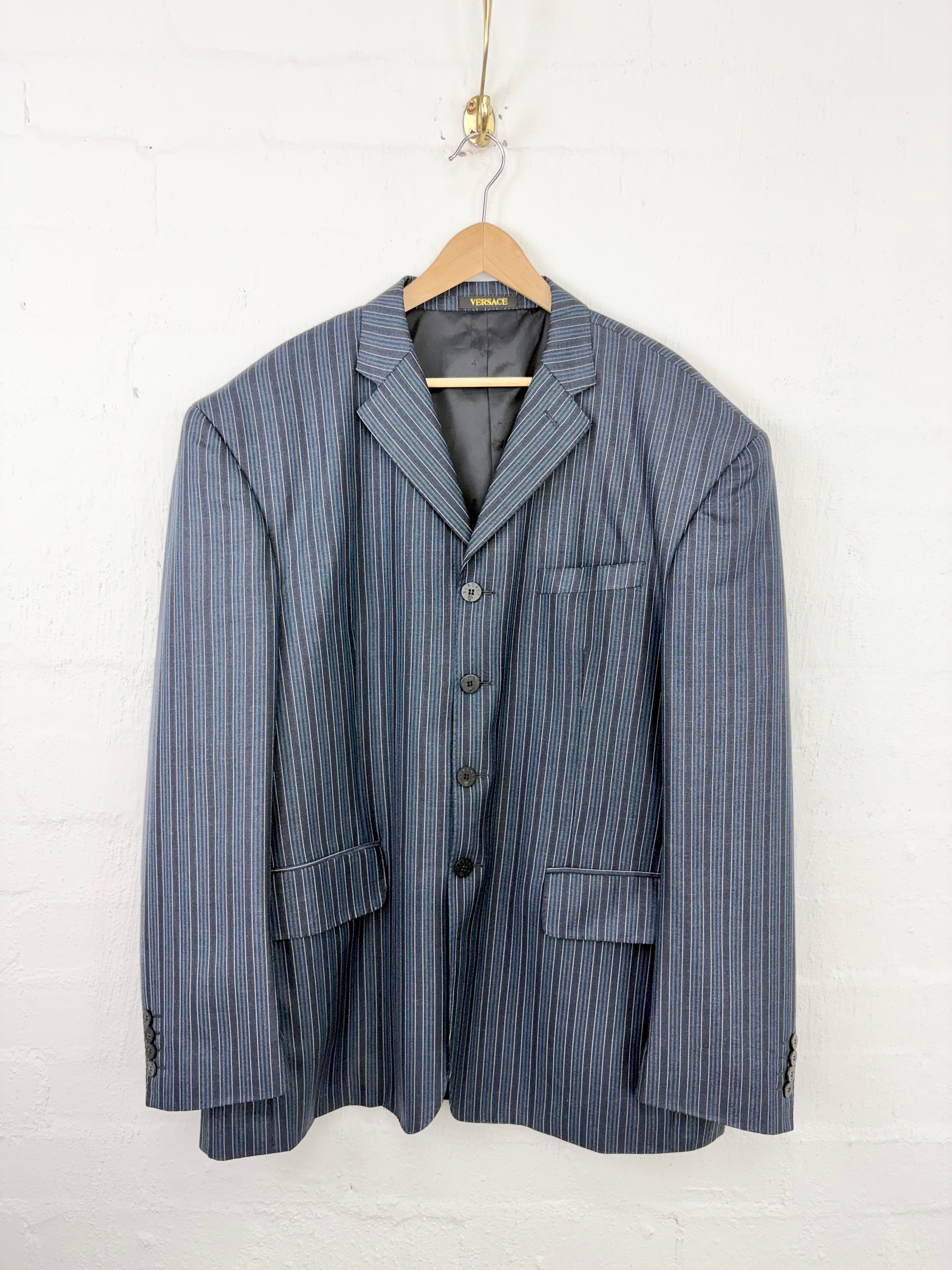 A men's pinstripe blazer jacket in blue and gray hanging on a wooden hanger against a white painted brick wall.
