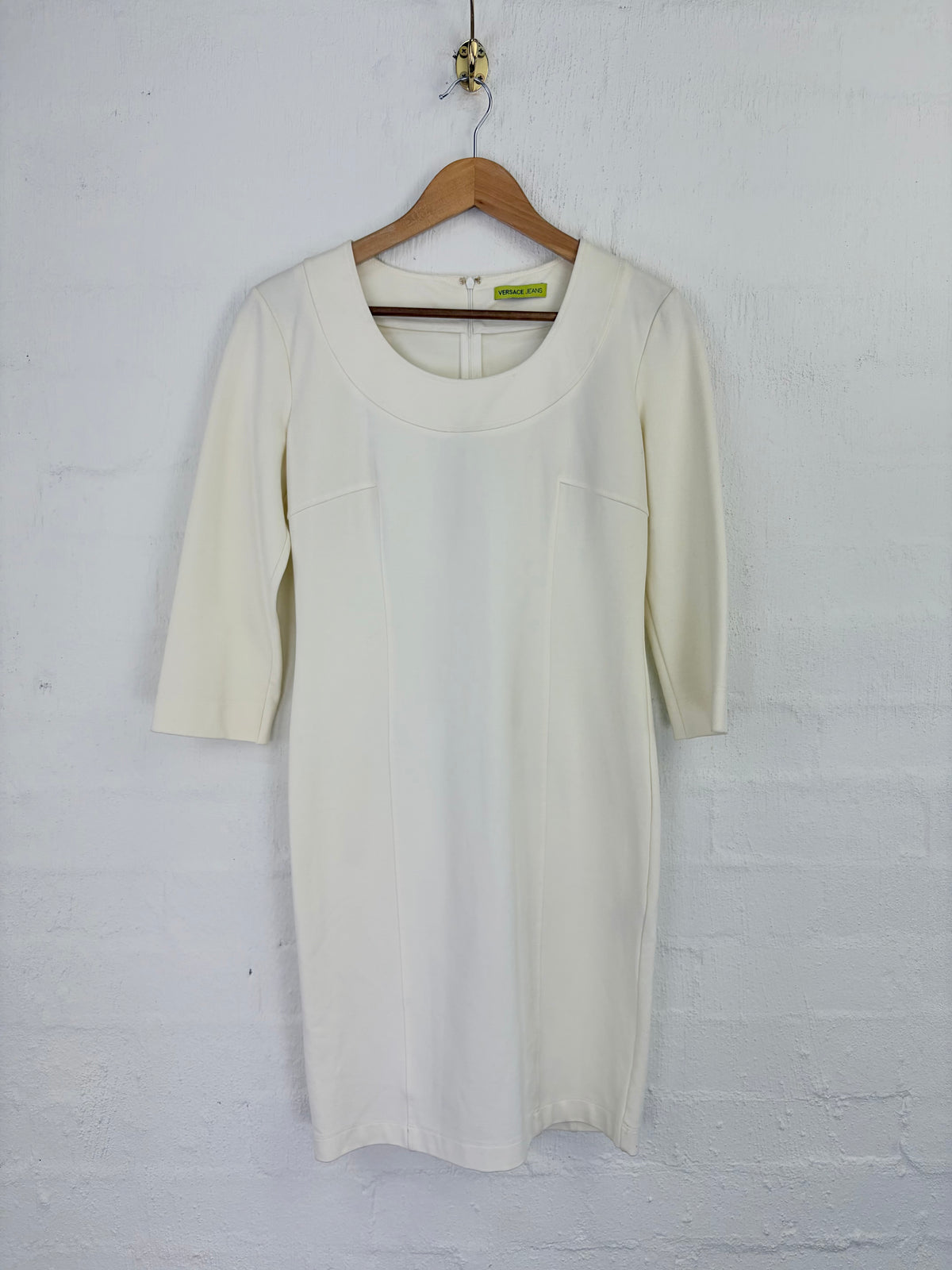 An off-white knee-length dress with three-quarter sleeves hanging on a wooden hanger against a white painted brick wall.