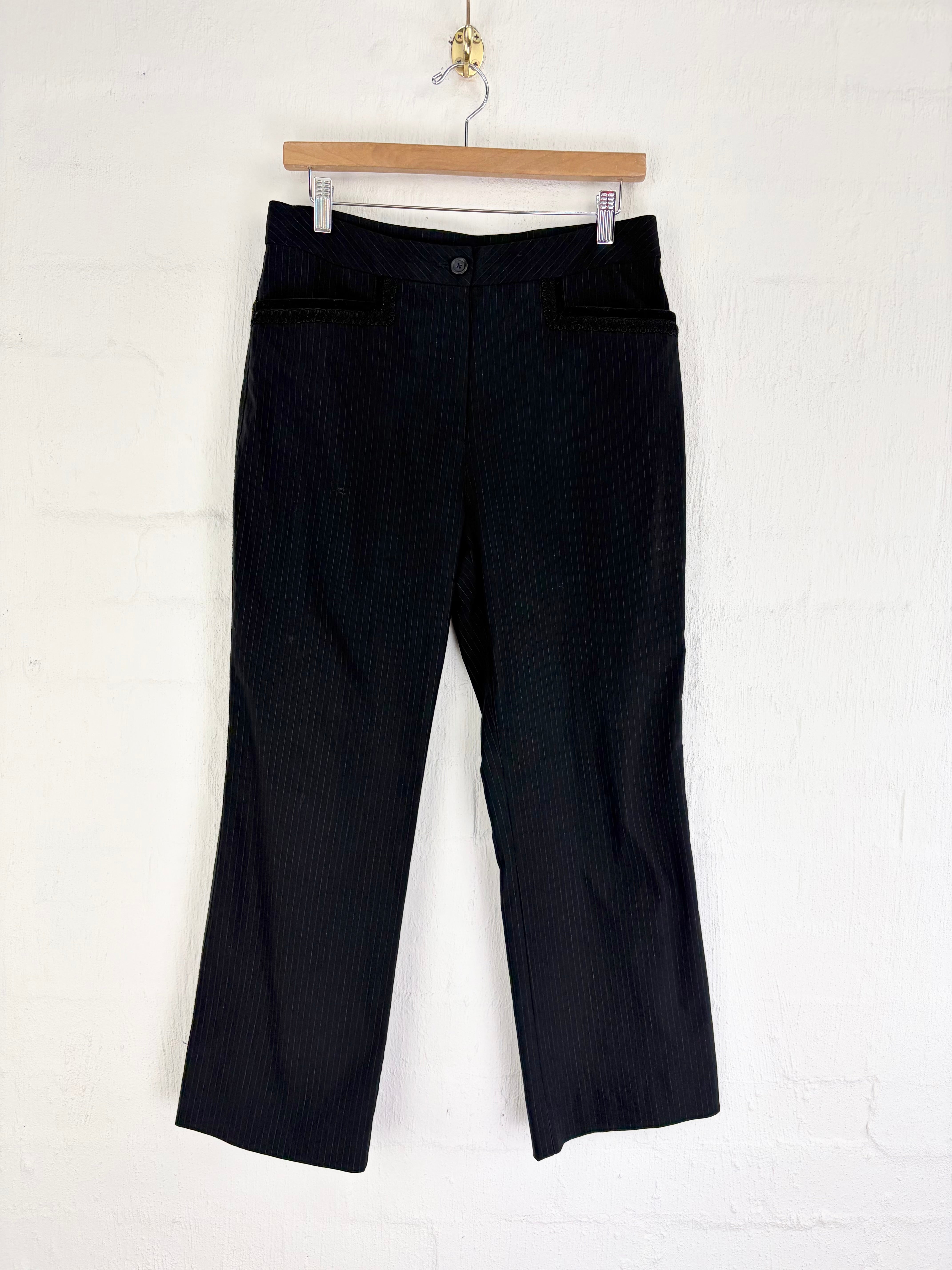 Black pinstriped cropped trousers hanging from a wooden hanger against a white brick wall.