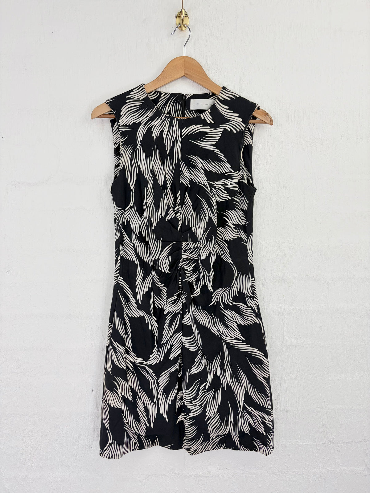 A sleeveless black and white patterned dress hanging on a wooden hanger against a white brick wall.
