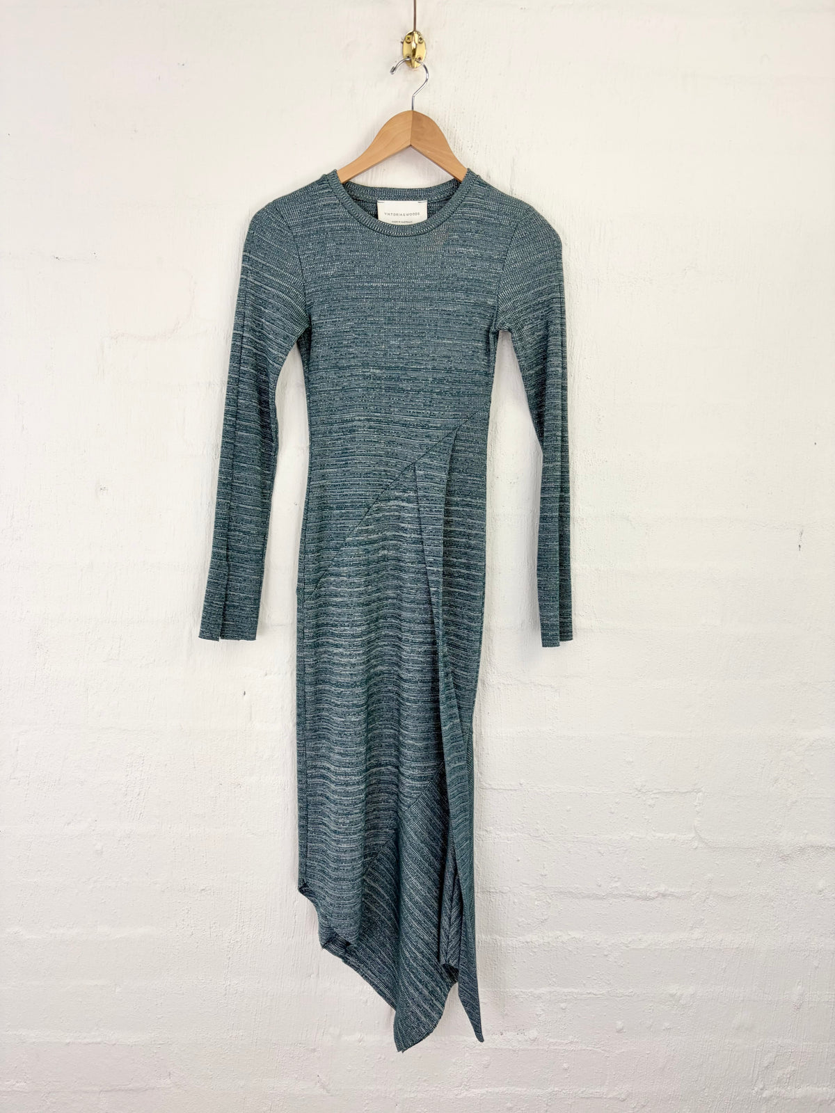 A long-sleeve knit dress in a heathered slate blue-gray color, hanging on a wooden hanger against a white painted brick wall.