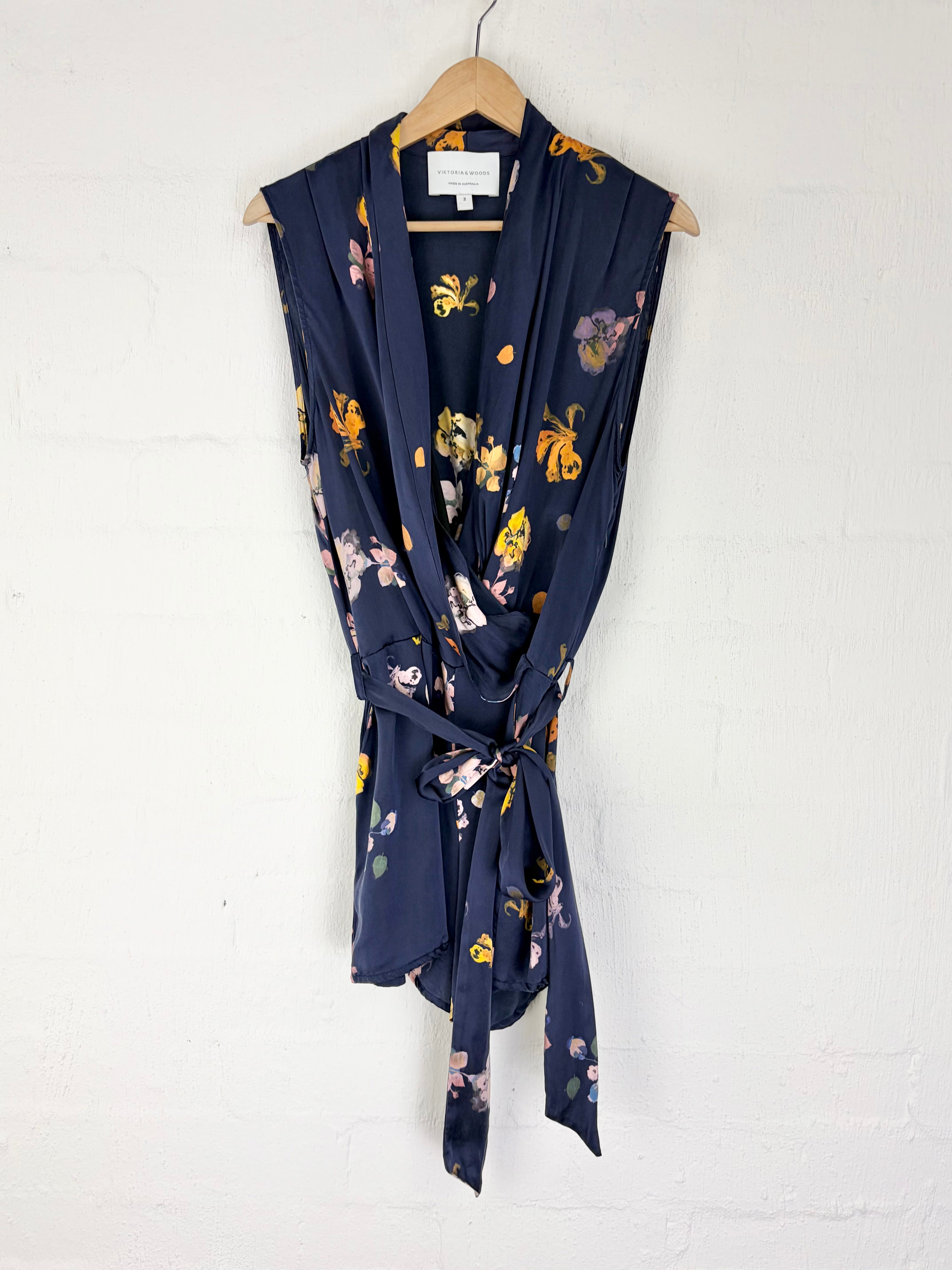 A navy blue sleeveless wrap-style top with a tie waist and a colorful floral print, hanging on a wooden hanger against a white painted brick wall.