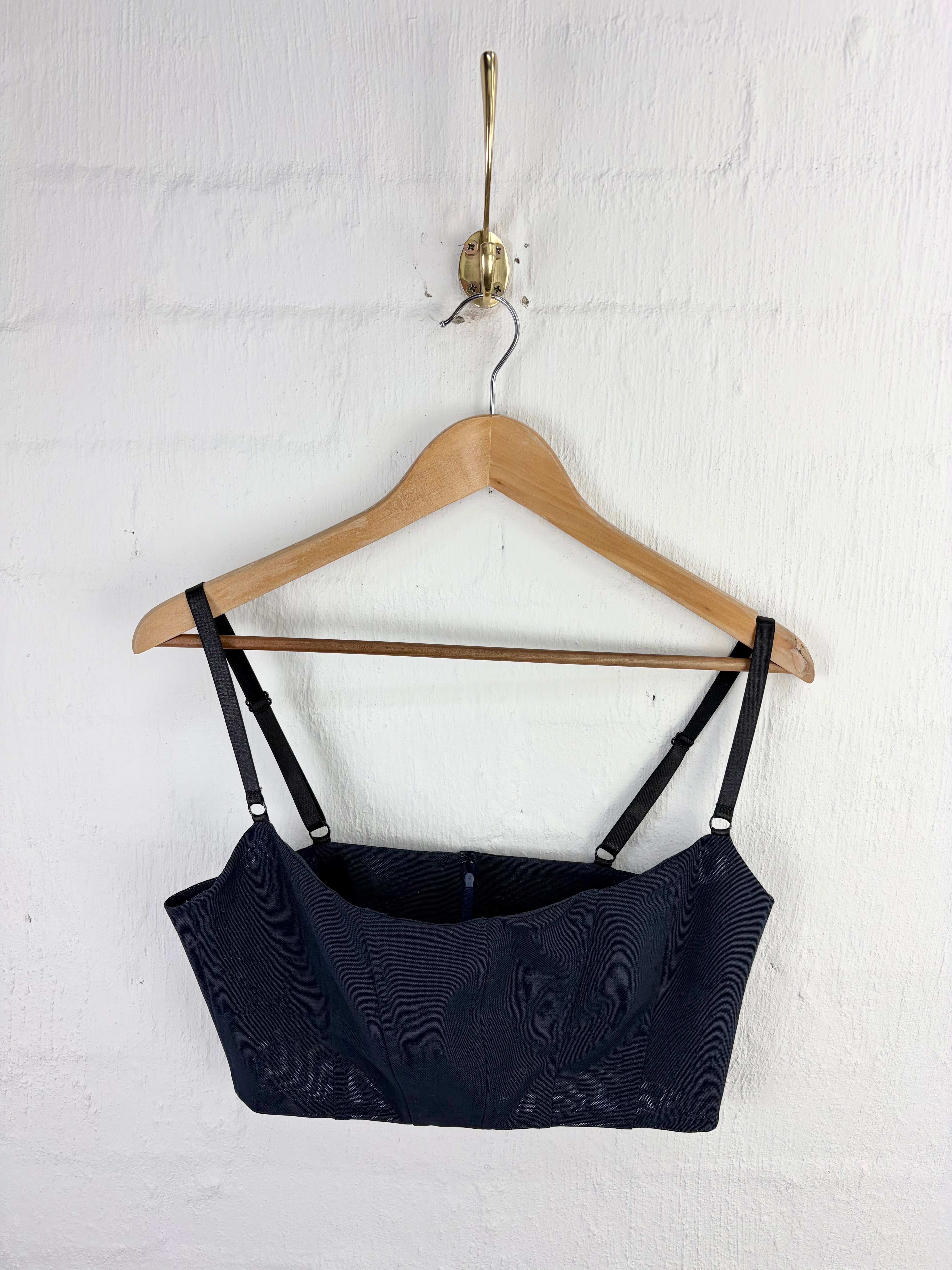 A dark navy blue camisole top with wide shoulder straps, hanging on a wooden hanger from a brass wall hook against a white painted brick wall.