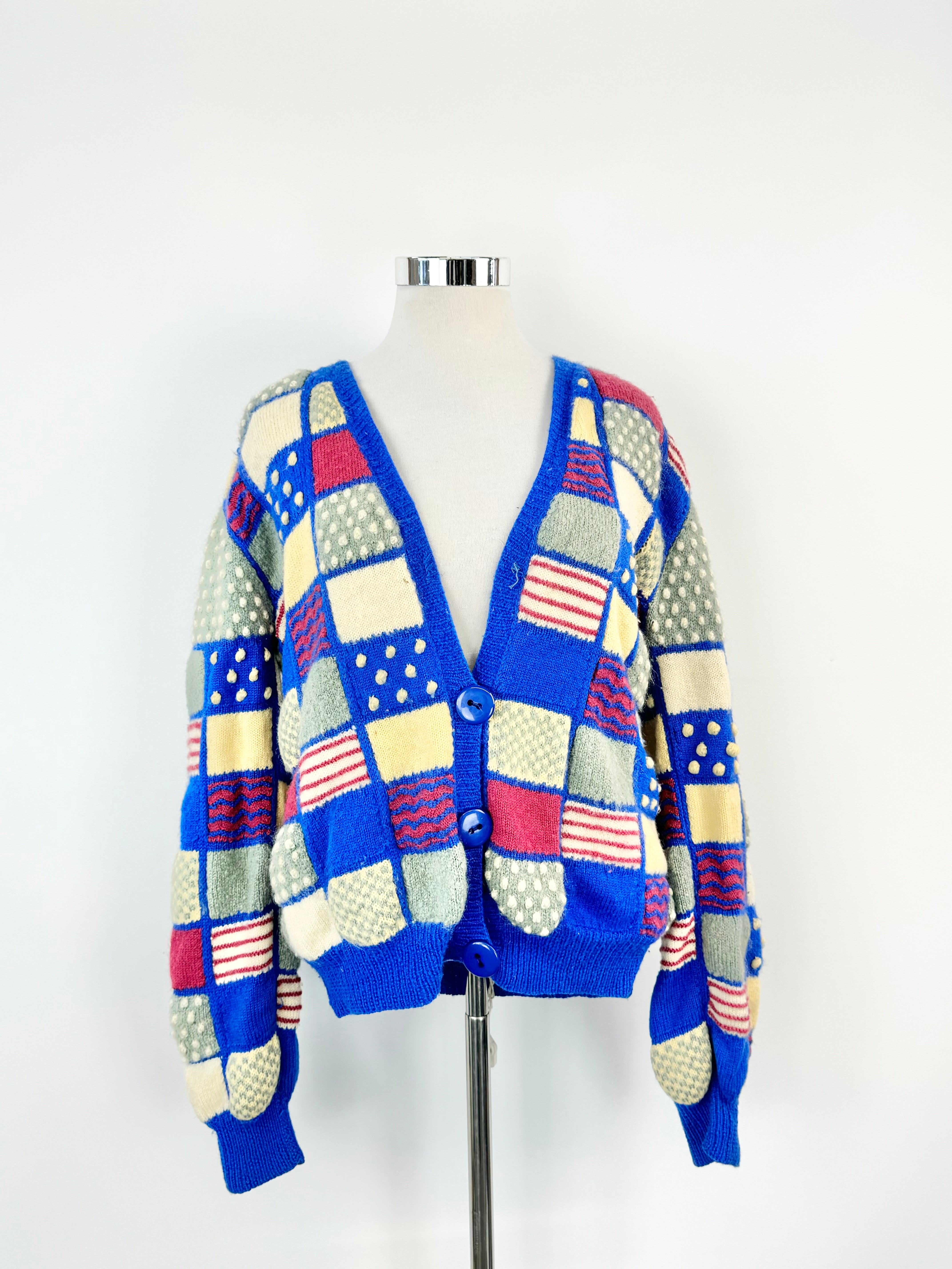 A vintage 70s blue, grey, and pink 3D knitted cardigan displayed on a mannequin against a white background.