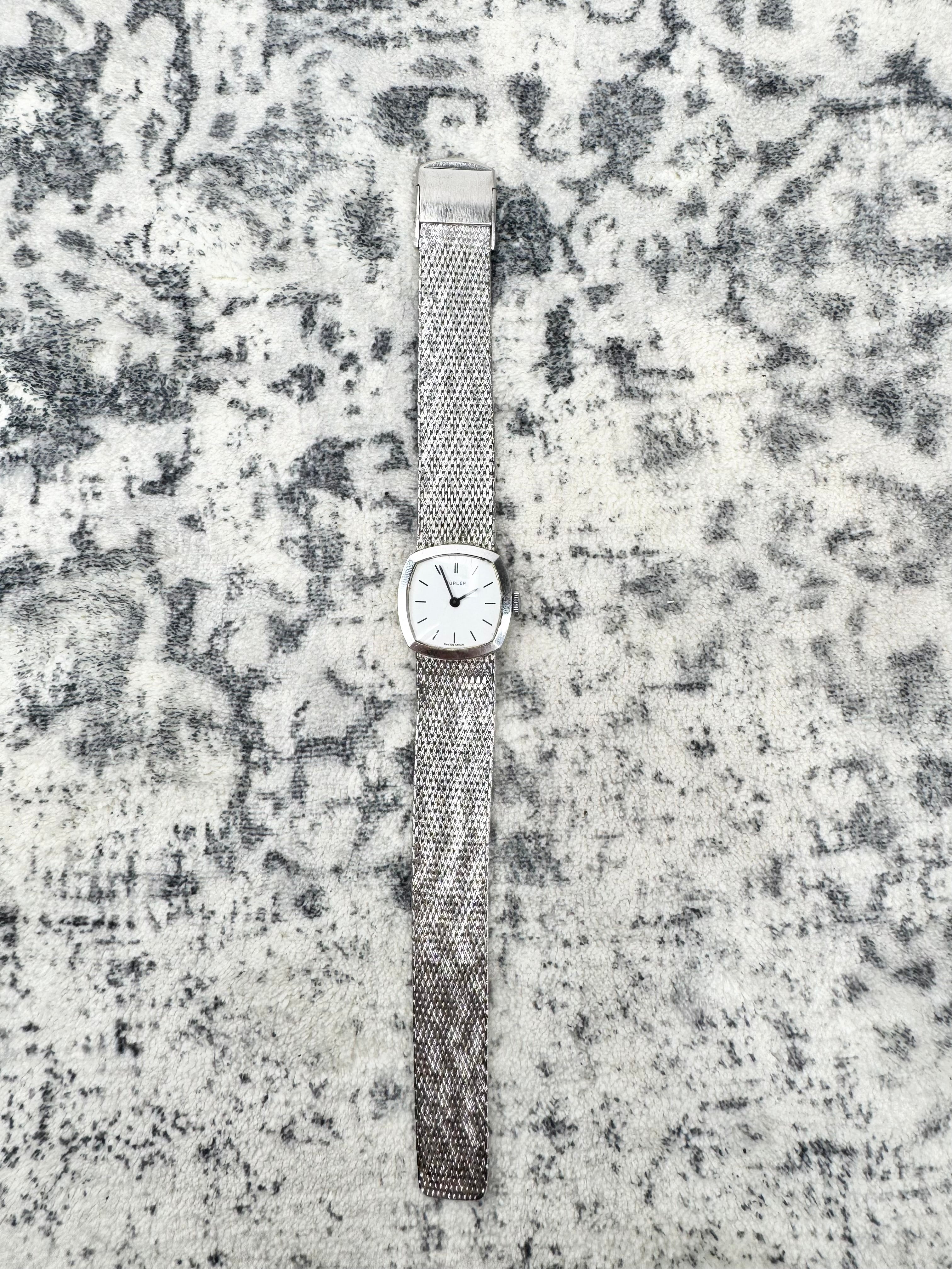 A vintage-style wristwatch with a silver-toned mesh band and a square white dial, laid flat on a textured gray and white surface.