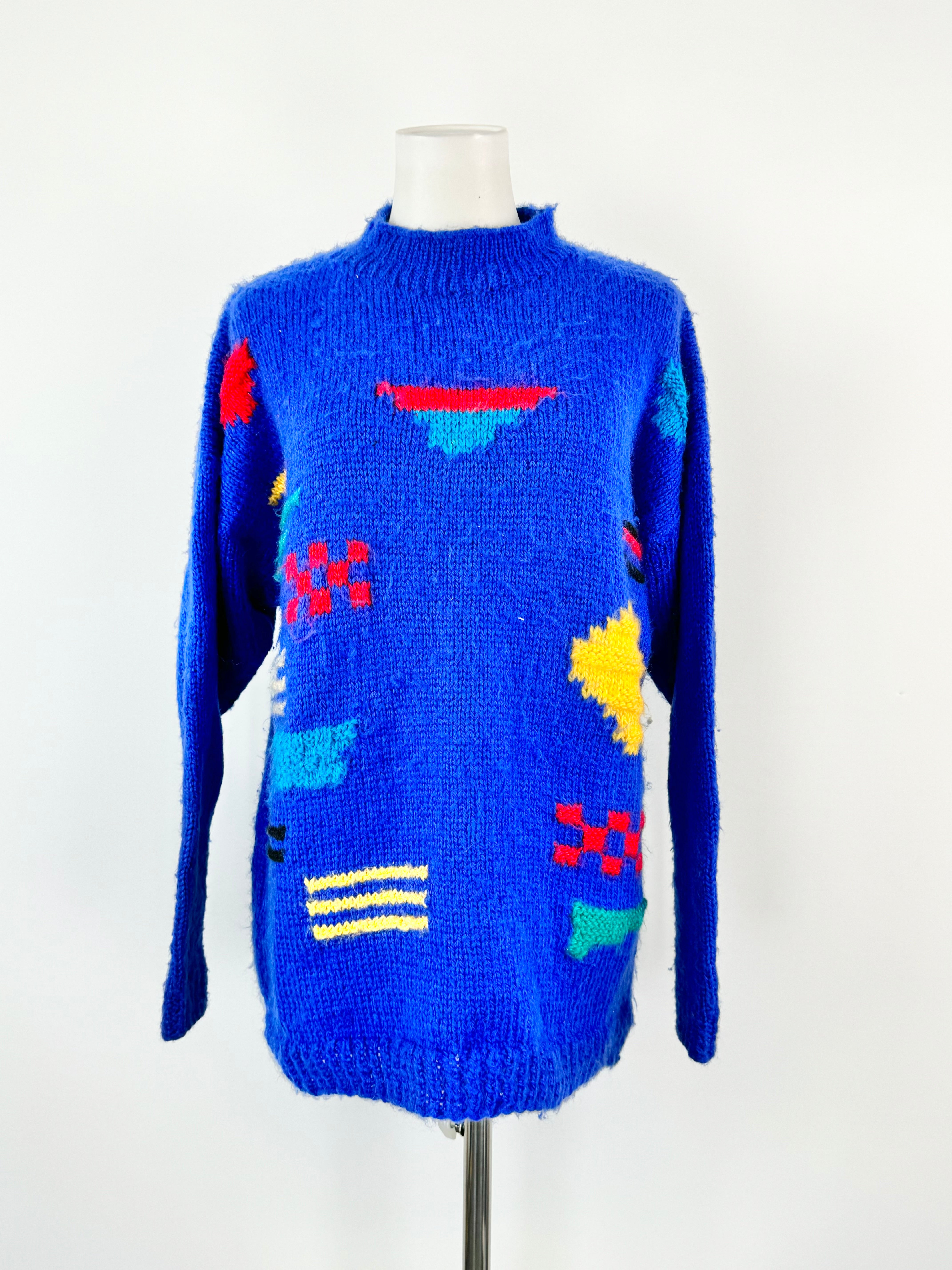 A bright electric blue hand knitted jumper with colorful geometric patterns on the front, displayed on a mannequin.