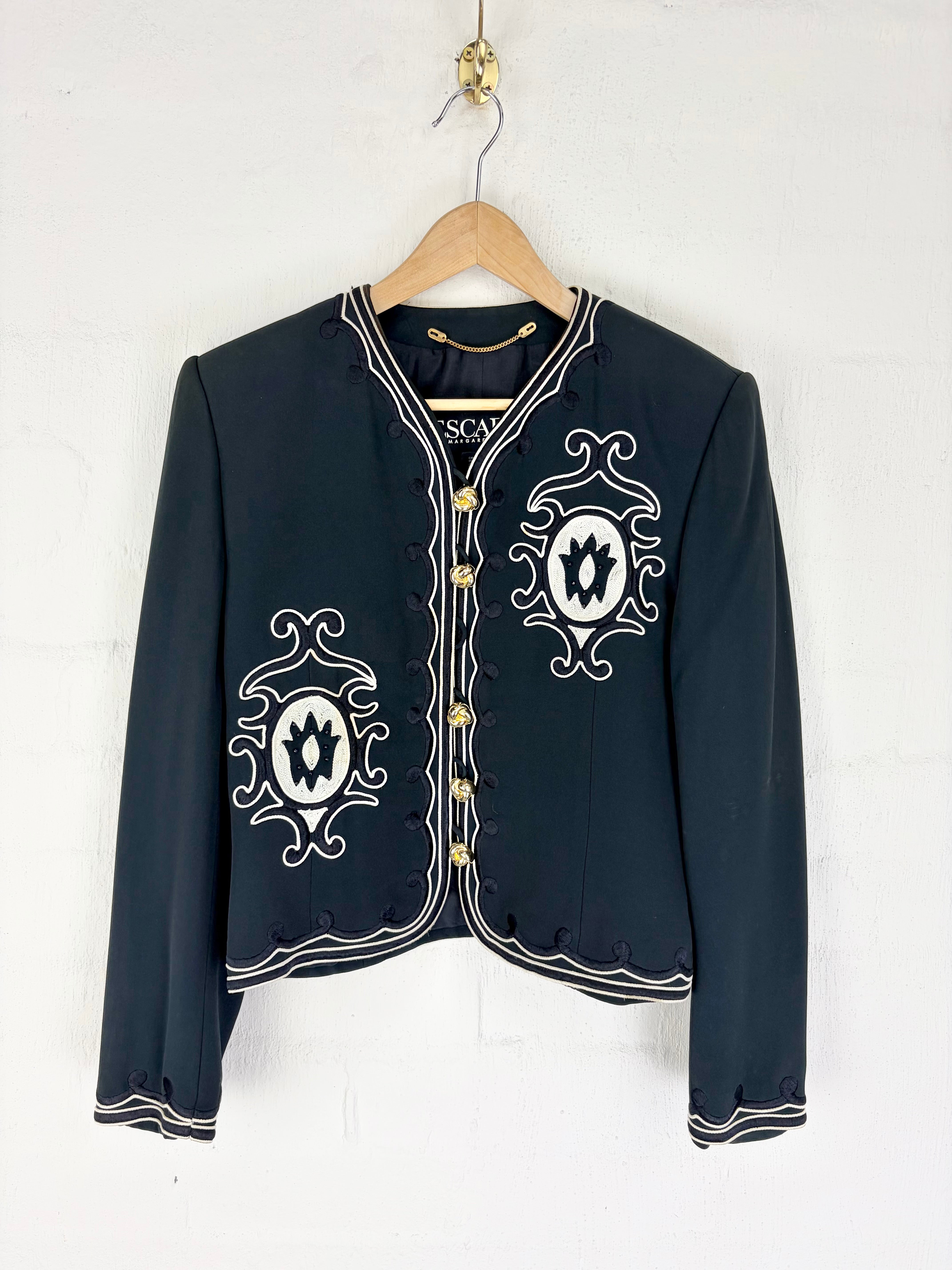 A dark navy blue cropped jacket with white embroidered patterns and gold buttons, hanging on a wooden hanger against a white wall.