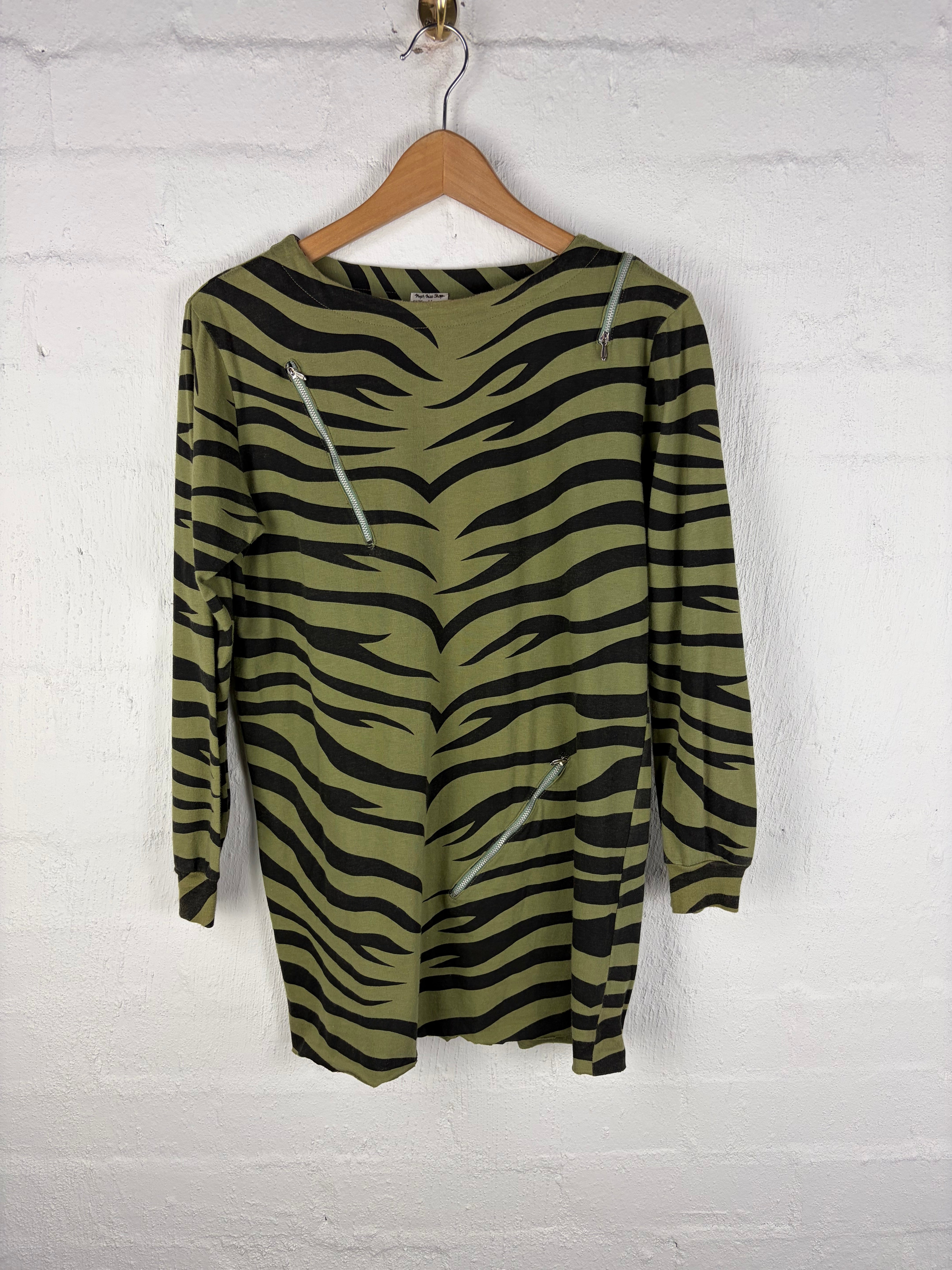 A long-sleeve knit top with an olive green and black zebra stripe pattern, draped on a wooden hanger against a white brick wall.