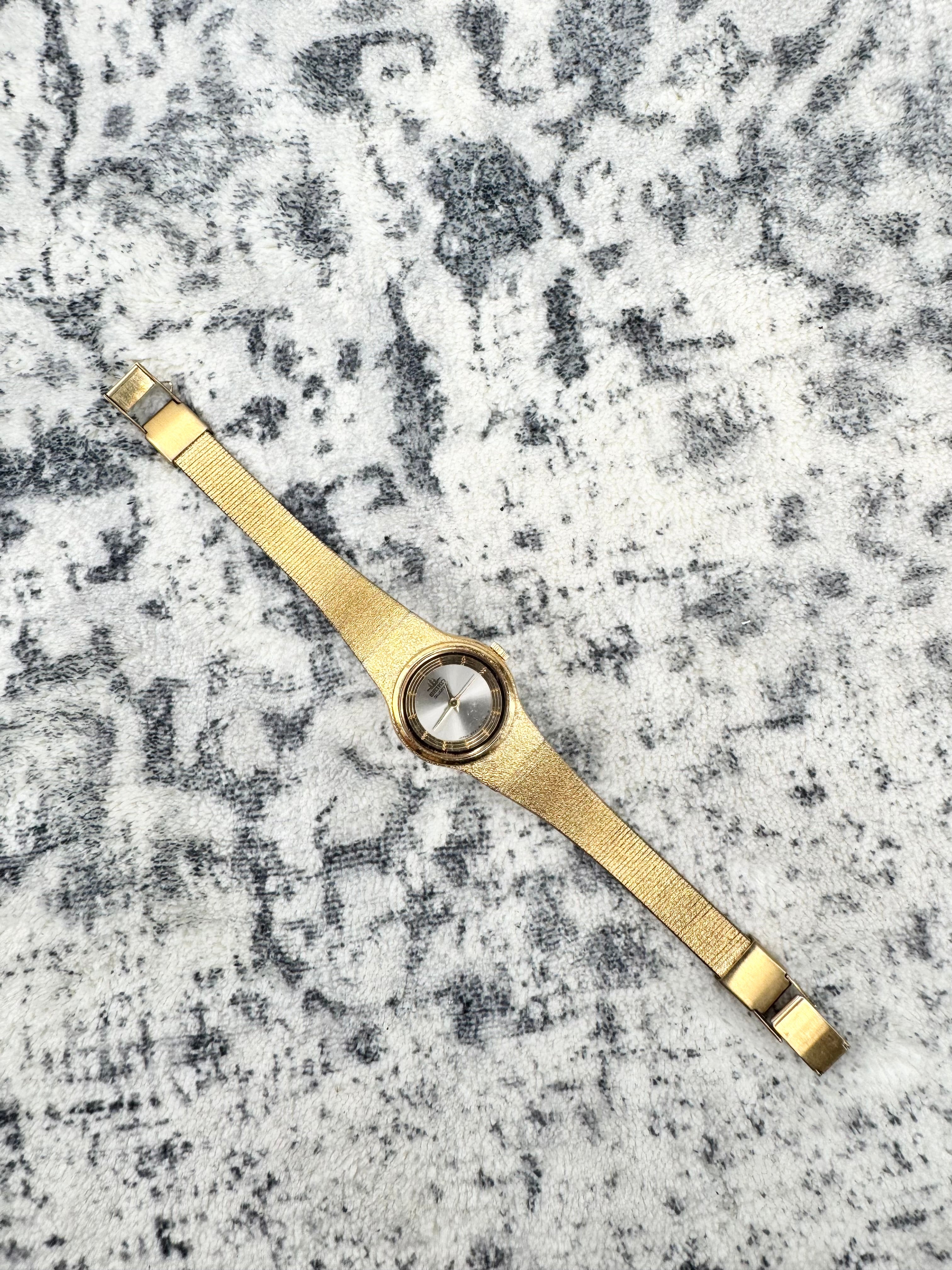 A gold-tone stainless steel watch band with a central circular dial, laid flat on a textured gray and white patterned rug.