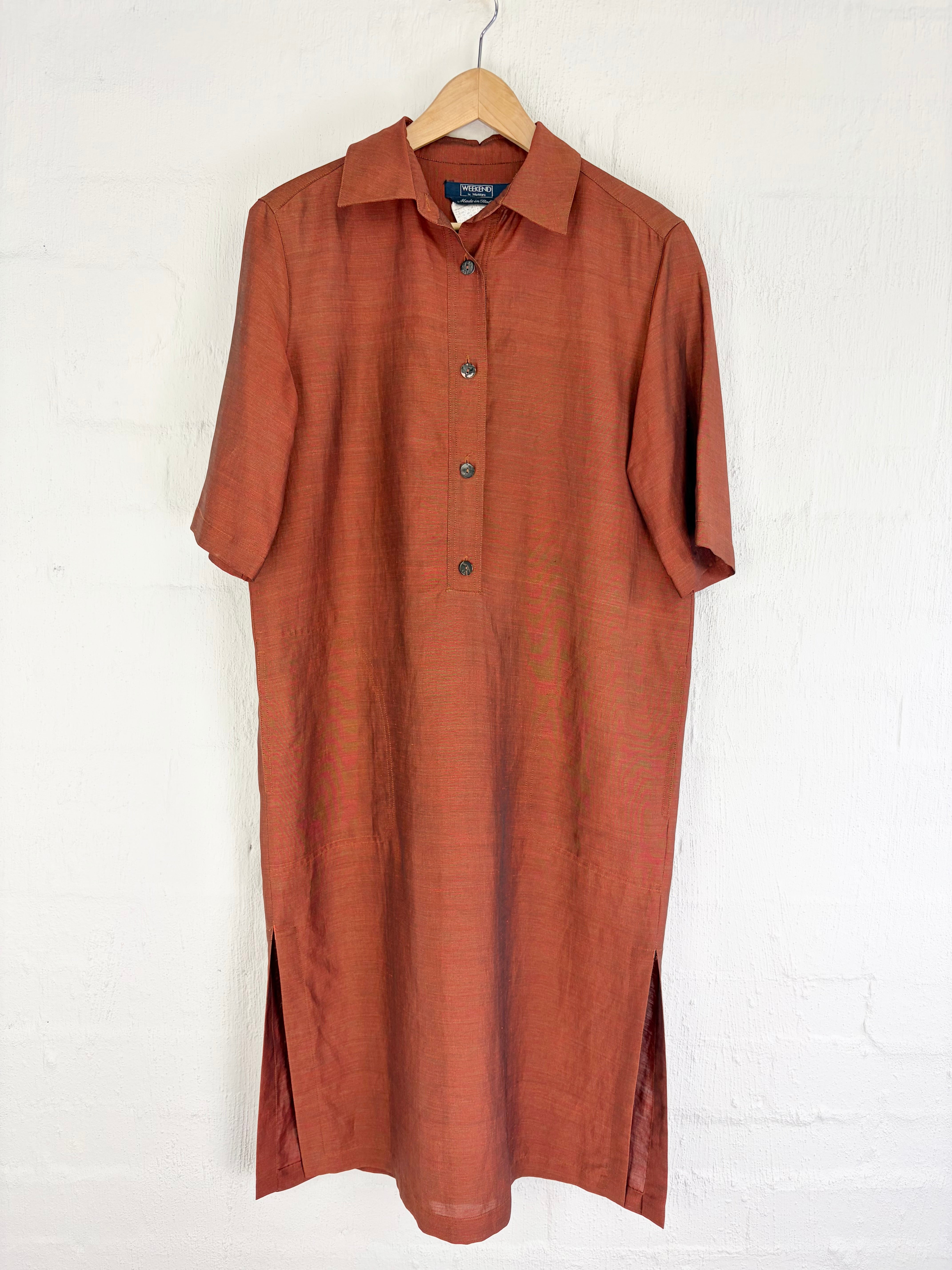 A short-sleeve collared button-front dress in a warm rust or terracotta color, hanging on a wooden hanger against a white painted brick wall.