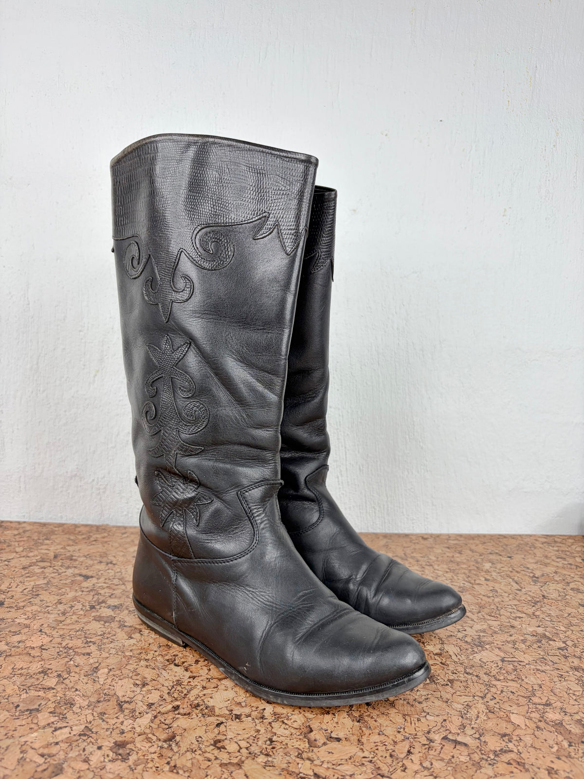 A pair of black leather mid-calf boots with western-style embroidery on the shaft, displayed on a brown textured surface against a plain off-white wall.