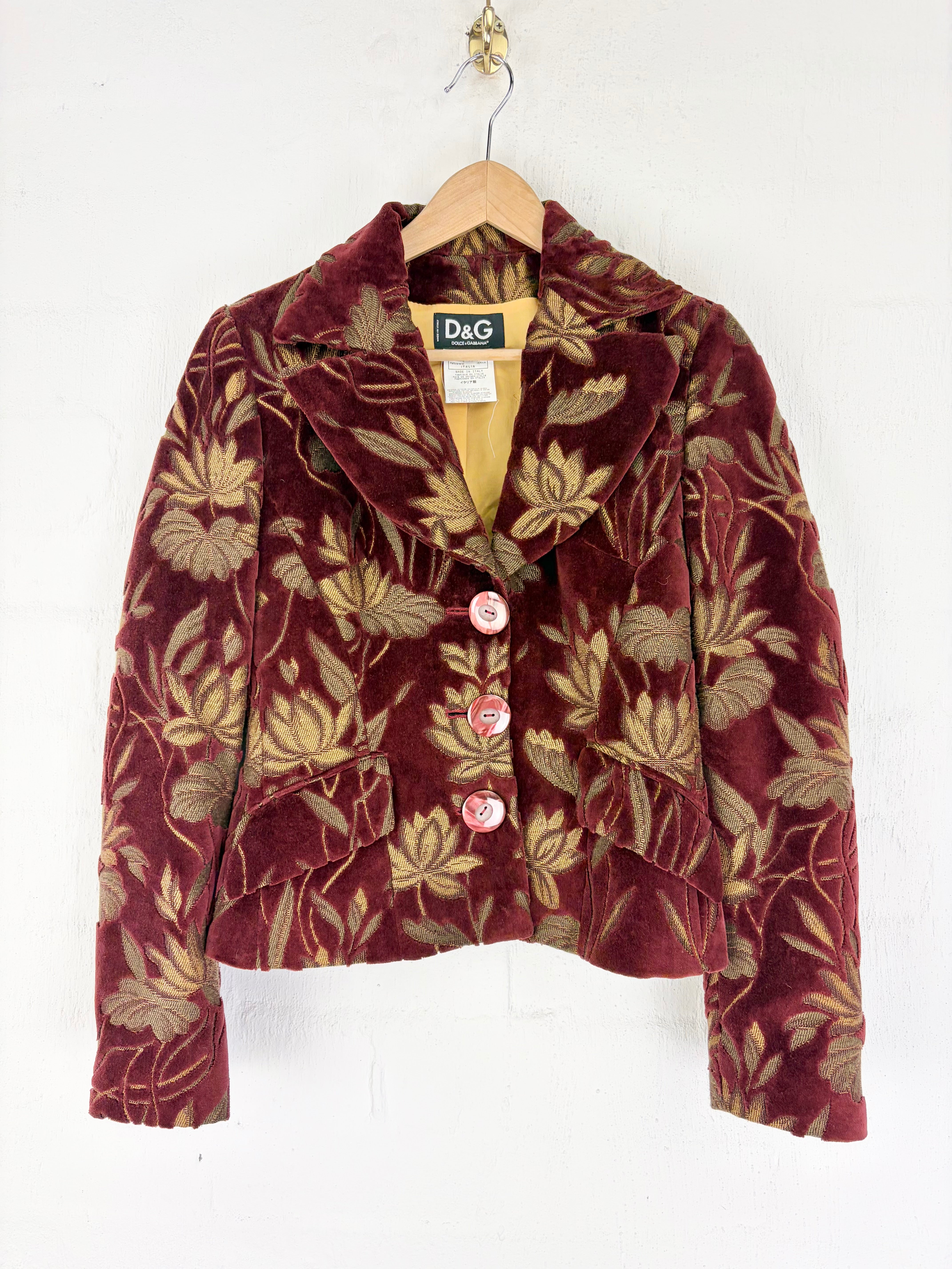 A vintage D&G floral velvet jacket in deep burgundy and gold, hanging on a wooden hanger against a white wall.