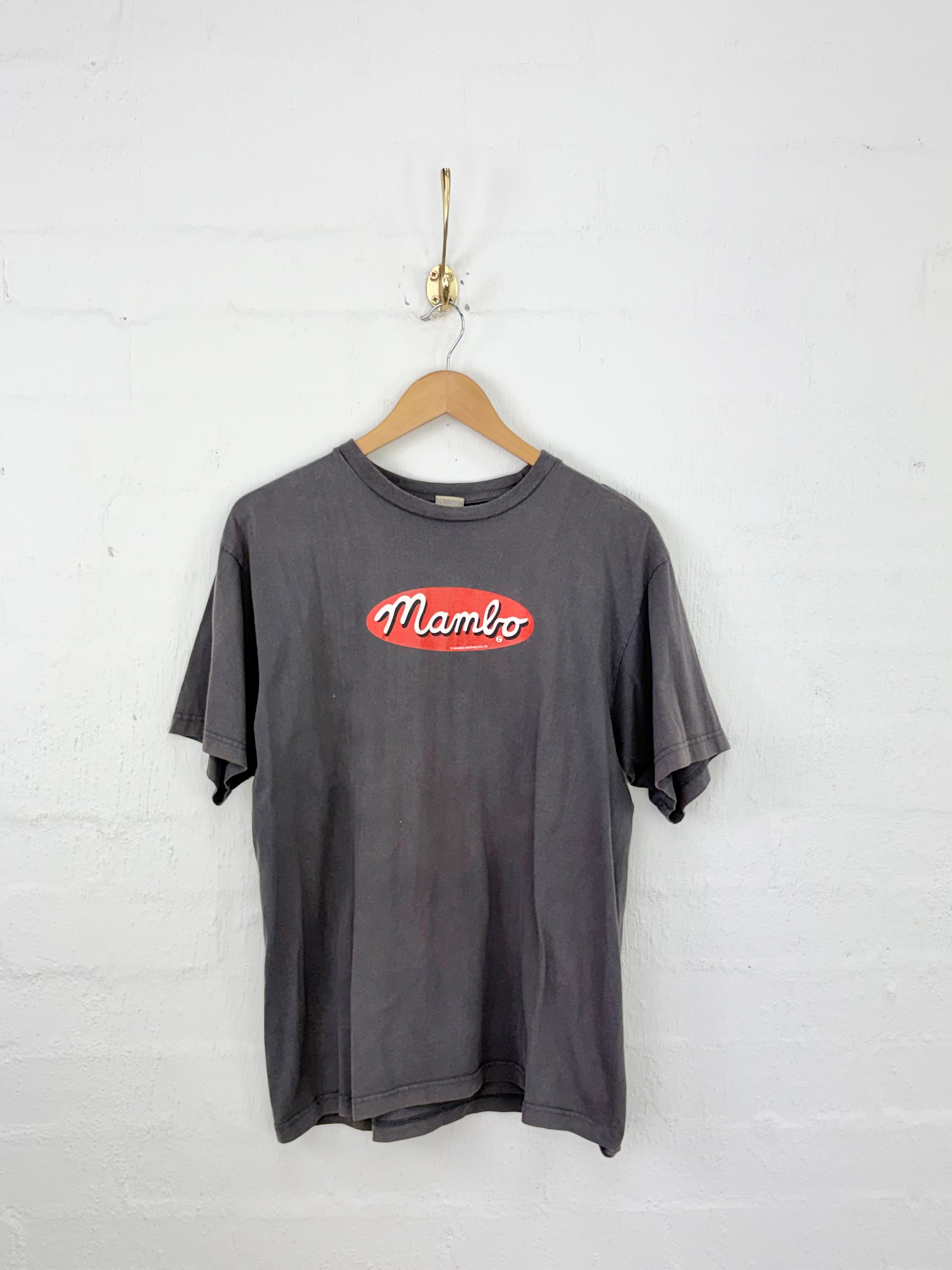 A faded dark gray short-sleeve t-shirt hanging on a wooden hanger against a white brick wall. The shirt features a red oval chest graphic with the word 'Mambo' in white script.