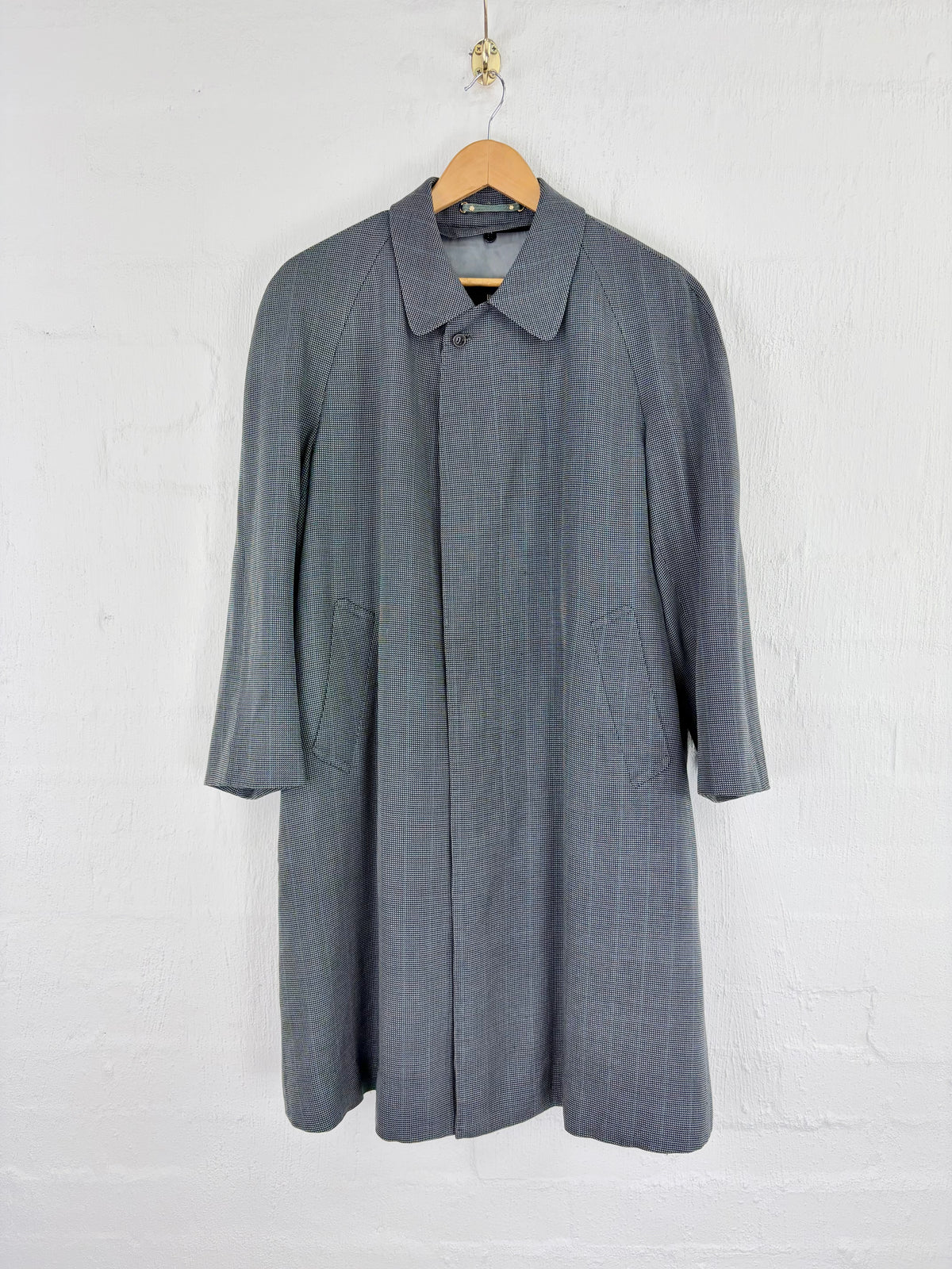 A gray textured coat with a pointed collar and three-quarter sleeves, hanging on a wooden hanger against a white brick wall.