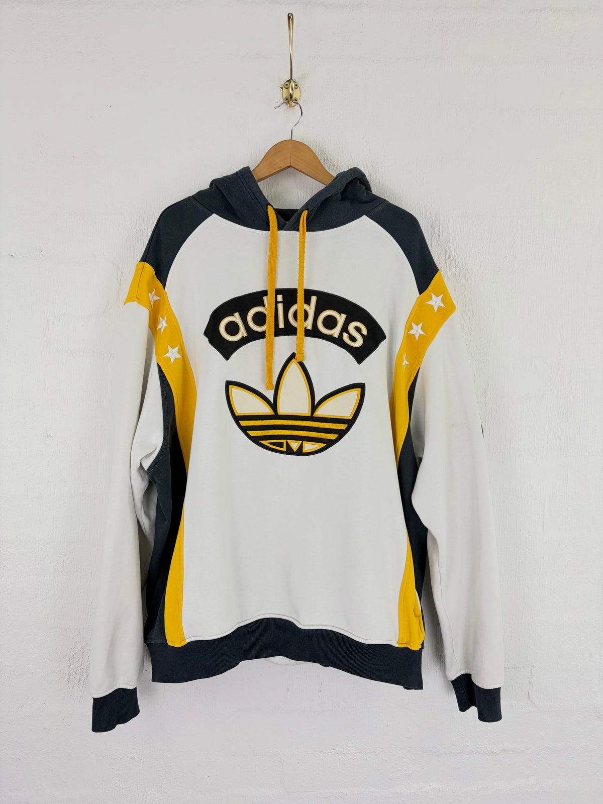 A white Adidas pullover hoodie with navy and yellow color blocking, featuring the classic Trefoil logo and 'adidas' text on the chest, hanging on a wooden hanger against a white brick wall.