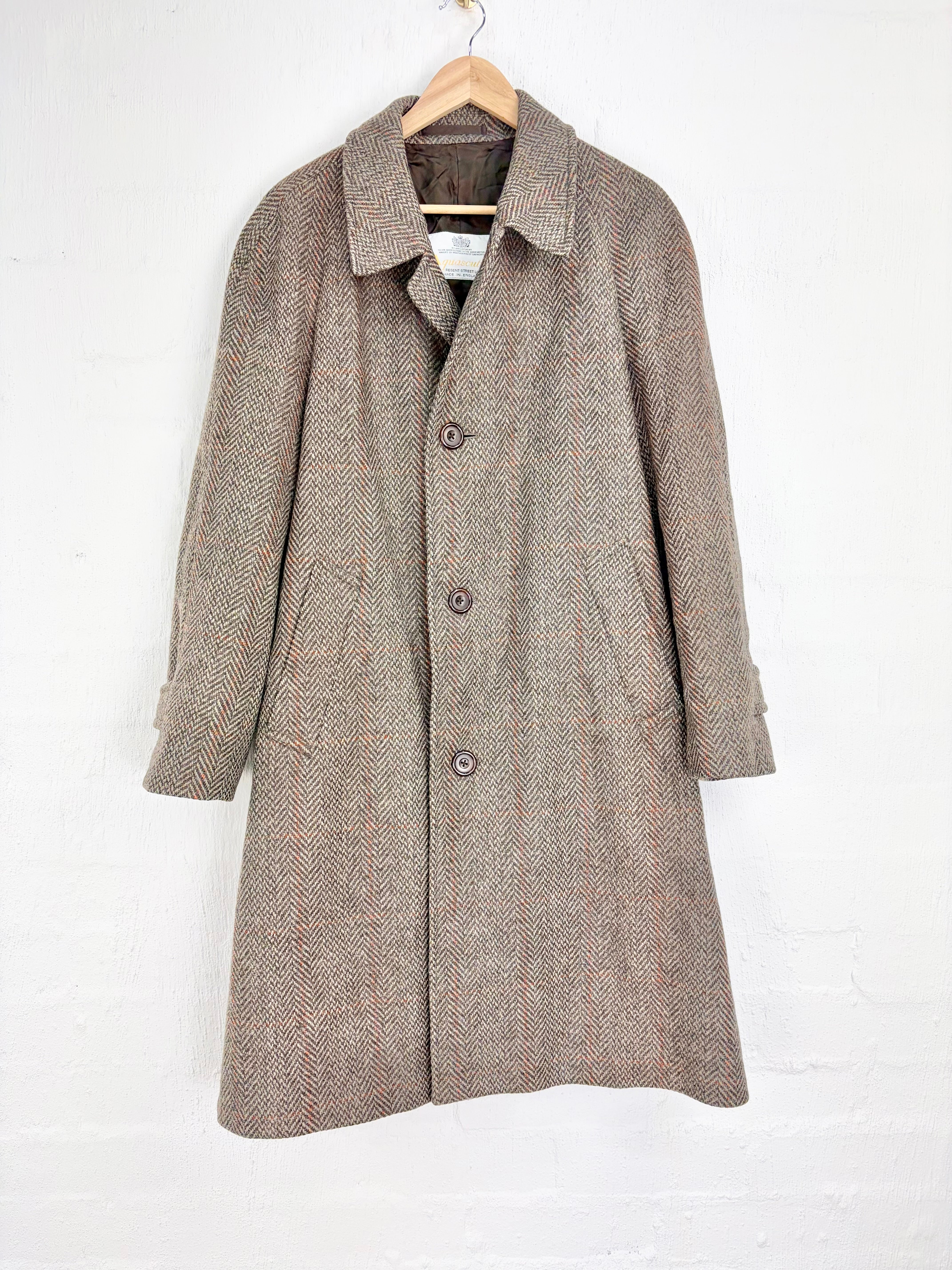 A mid-length herringbone coat in taupe and brown, hanging on a wooden hanger against a white painted brick wall.