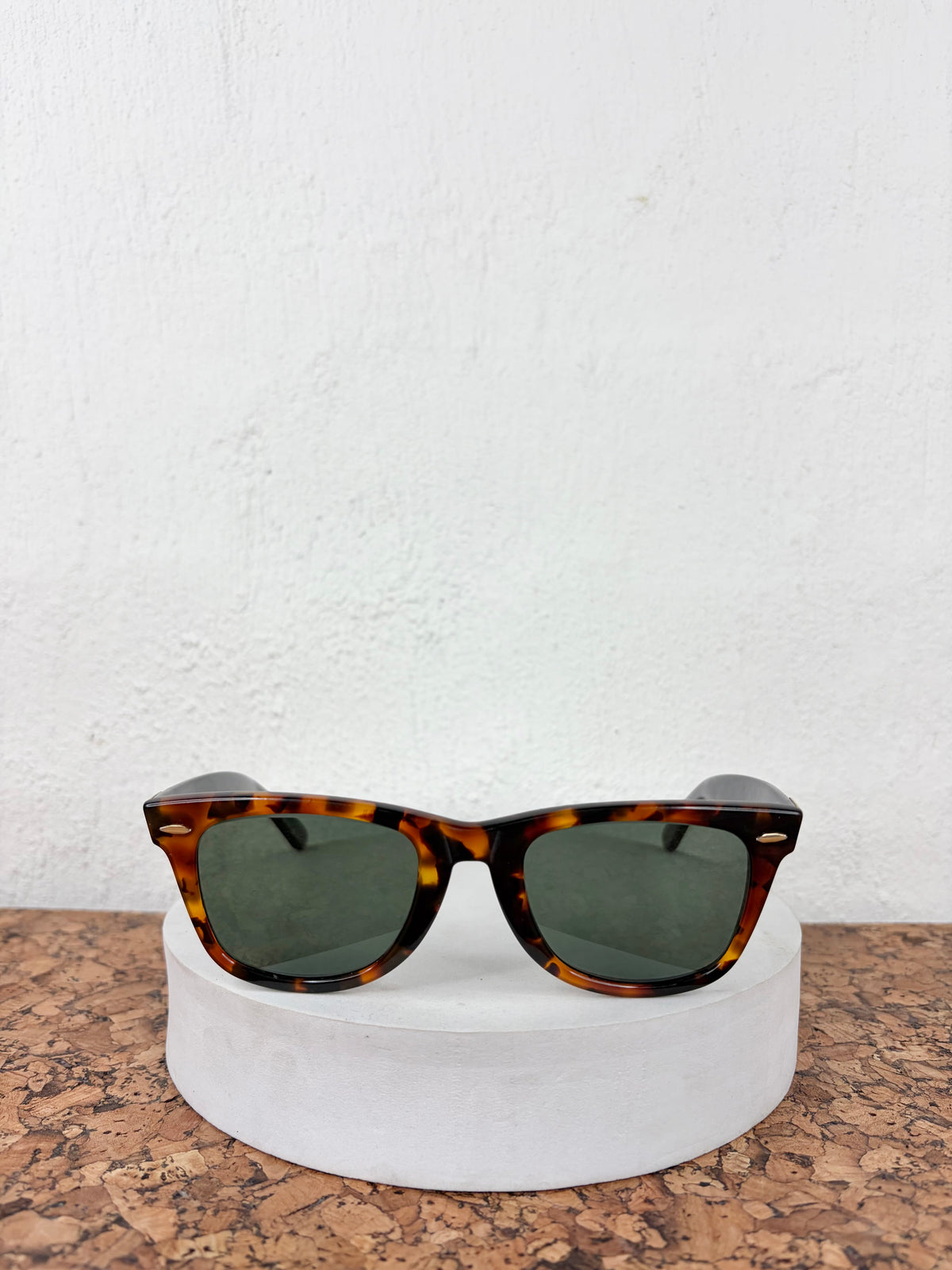 Tortoiseshell sunglasses with dark green lenses resting on a round white display stand against a white textured wall.