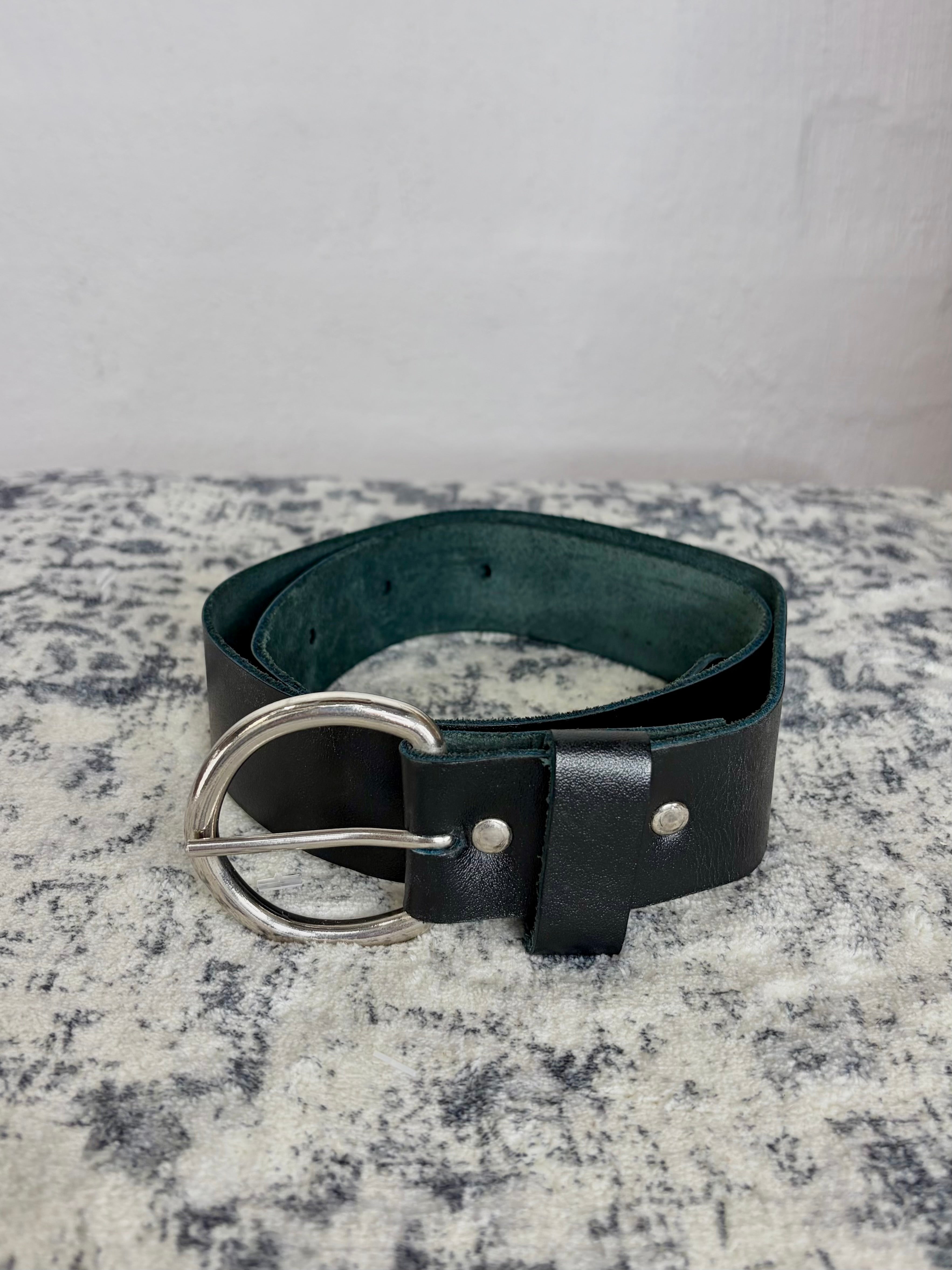 A black leather belt with a silver-tone metal buckle, displayed on a textured gray and white surface against a light gray background.