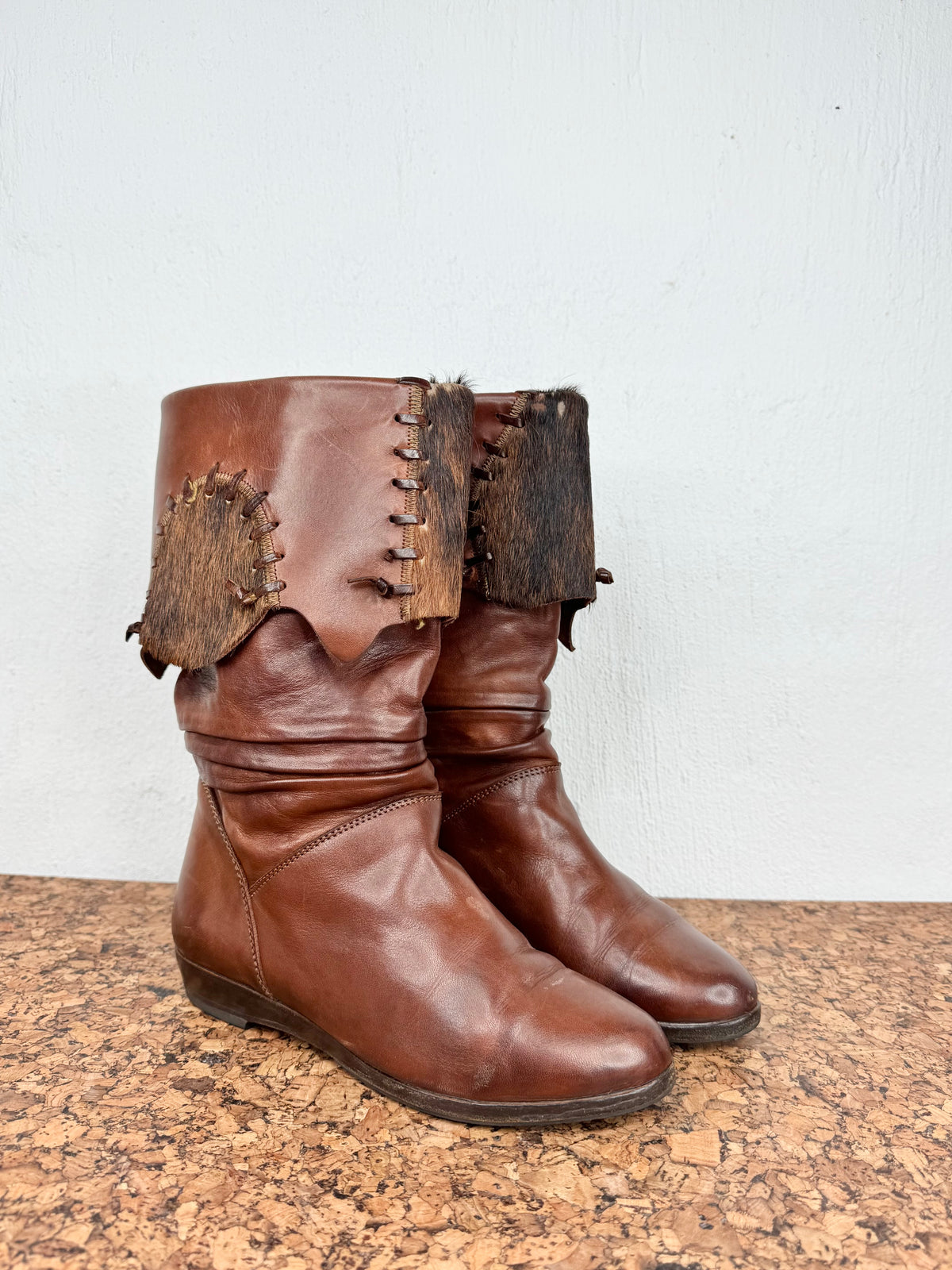 A pair of brown leather mid-calf boots with rustic animal-hide patches and decorative stitching on the shaft, displayed on a cork surface against a white wall.