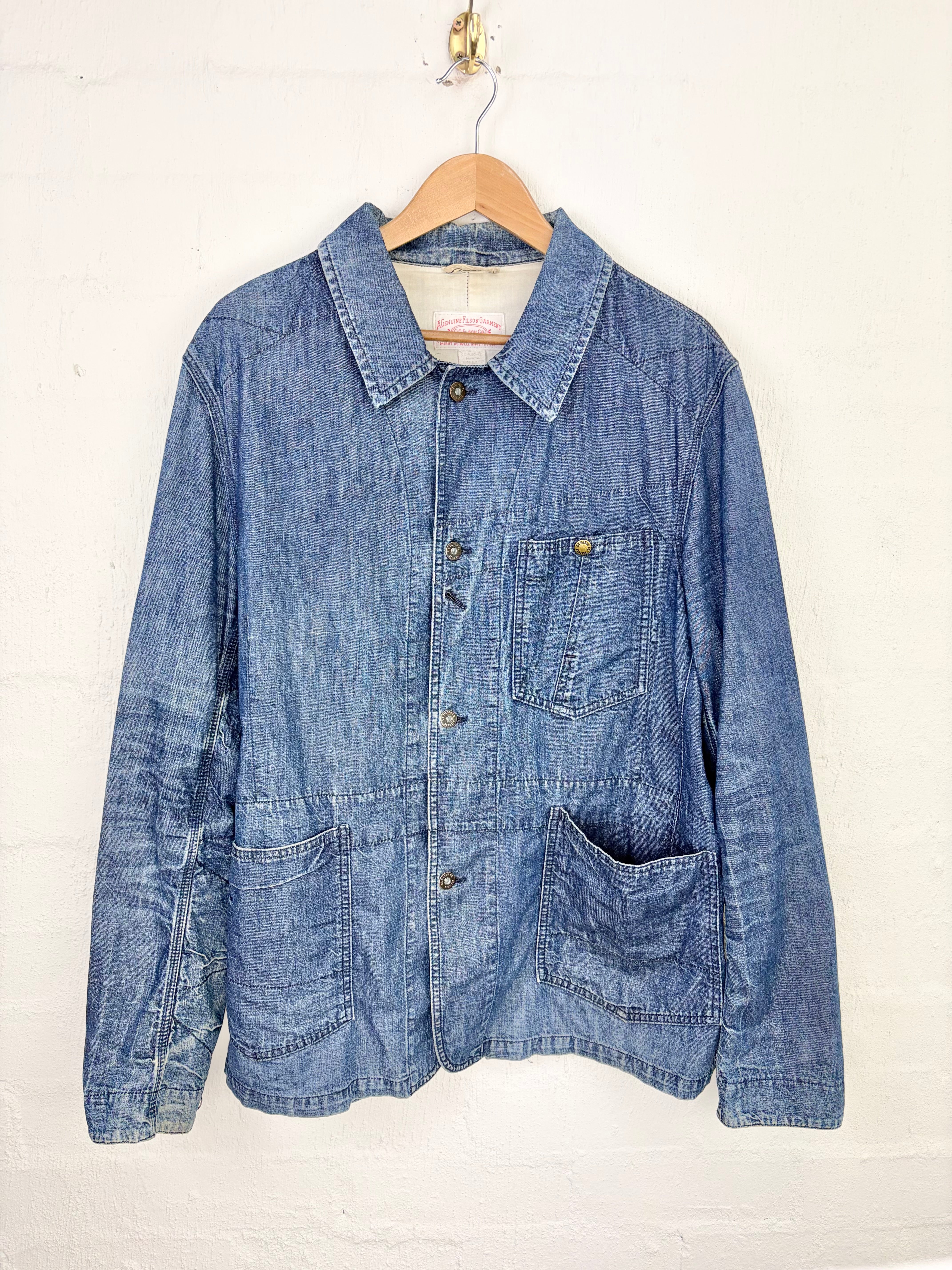 A blue denim work jacket hanging on a wooden hanger against a white painted brick wall. The jacket features a classic collar, button-up front, chest pocket, and two large patch pockets on the lower front.