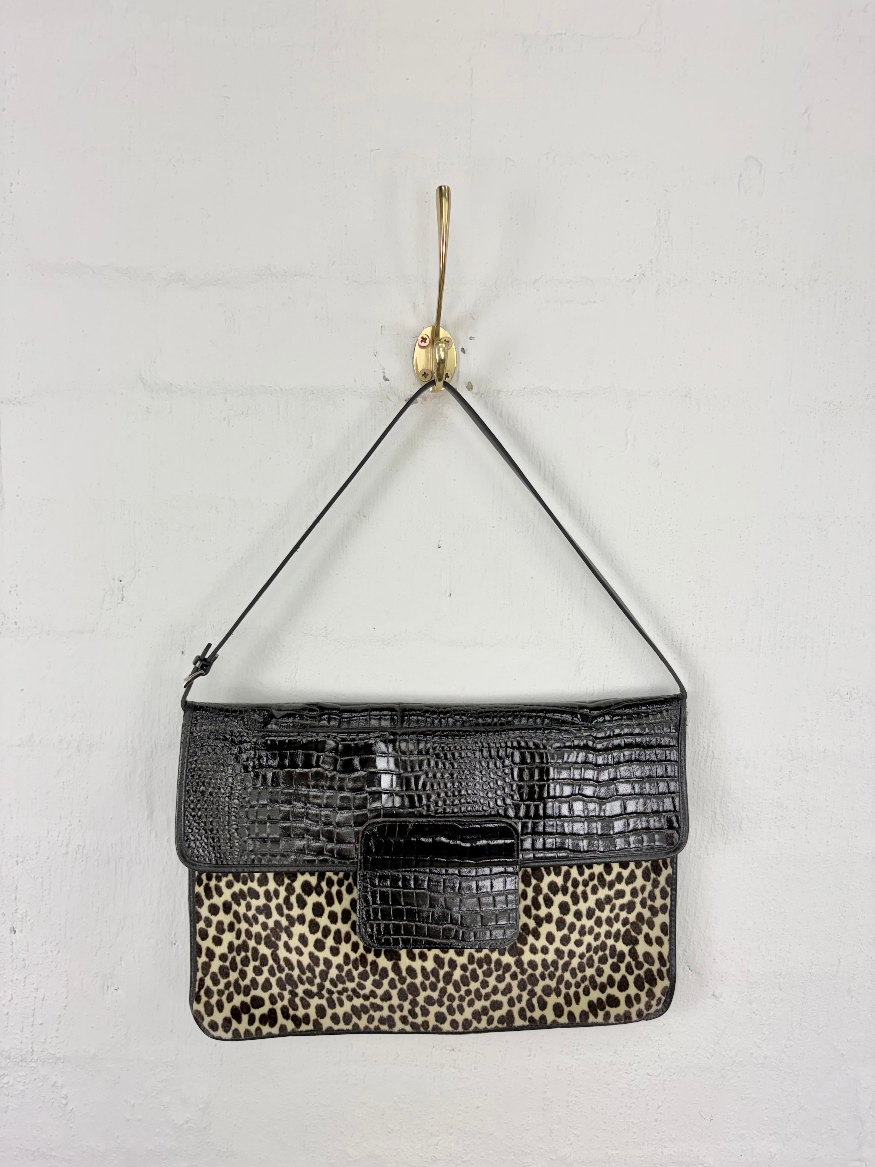 A rectangular shoulder bag with a black crocodile-embossed top flap and a leopard-print lower body, hanging from a gold hook against a white wall.
