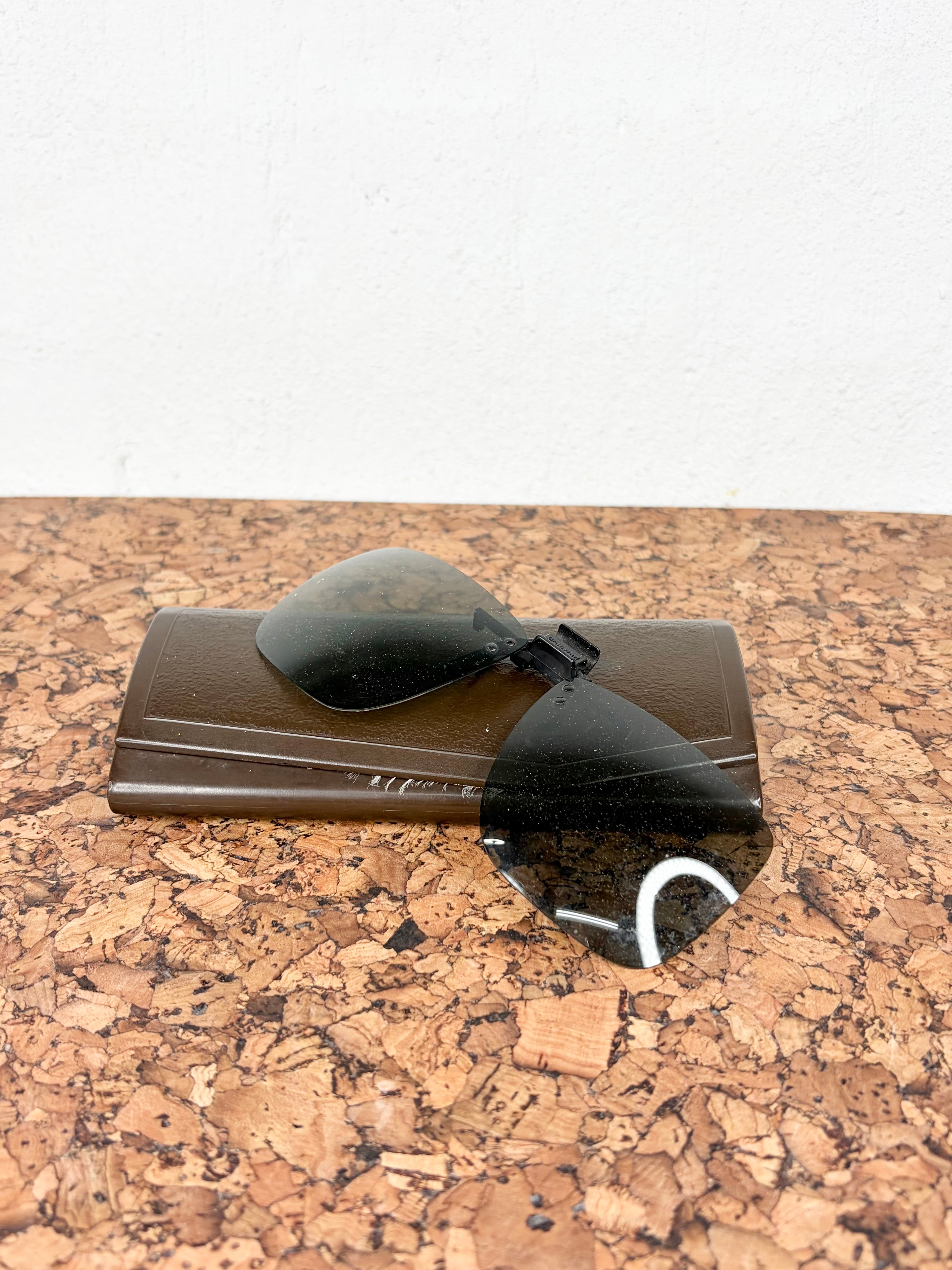 A pair of dark tinted clip-on sunglasses lenses resting on a brown rectangular case, placed on a cork-textured surface against a white wall.