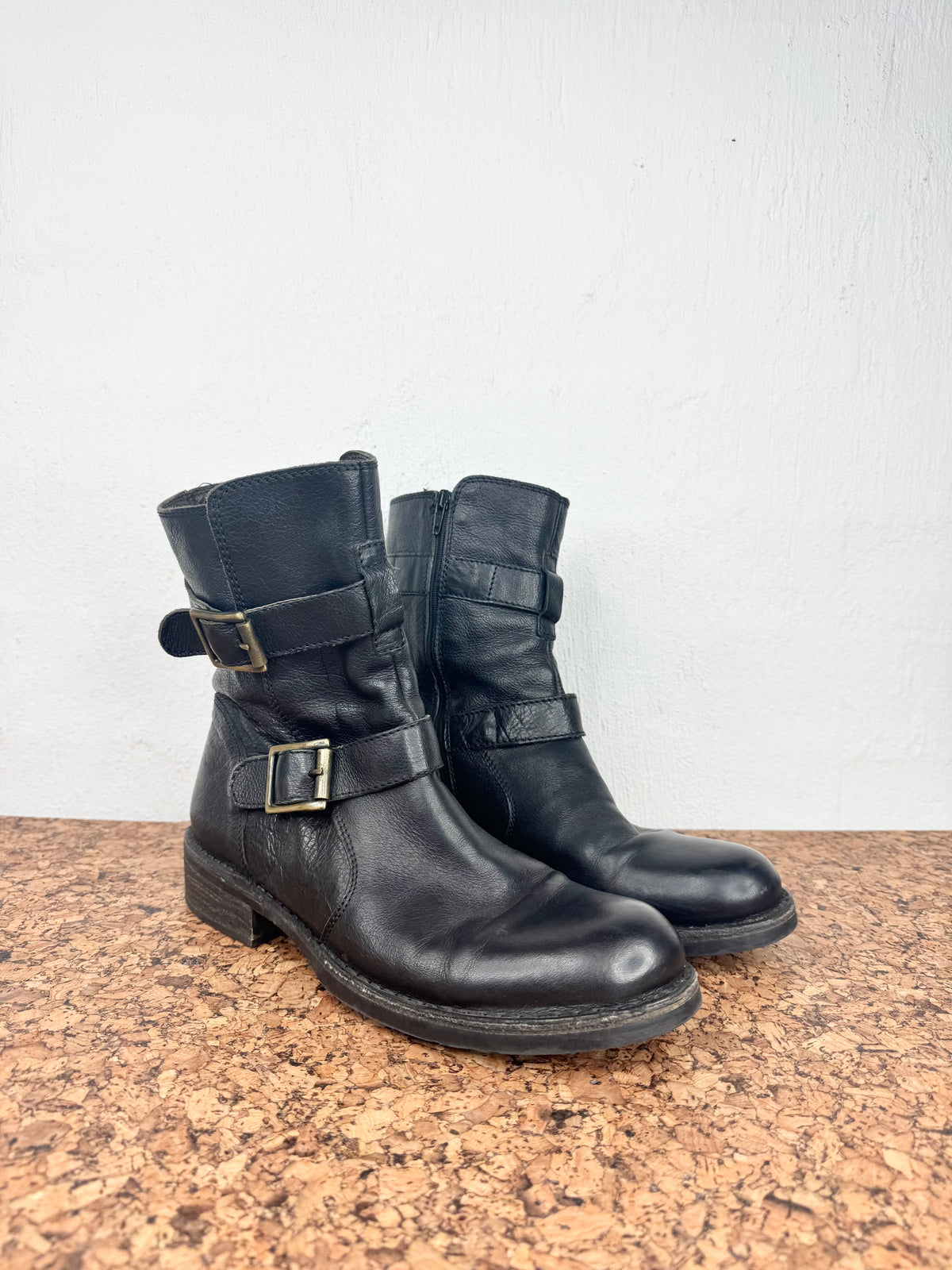 A pair of black leather ankle boots with buckle straps, resting on a brown speckled countertop against a white wall.