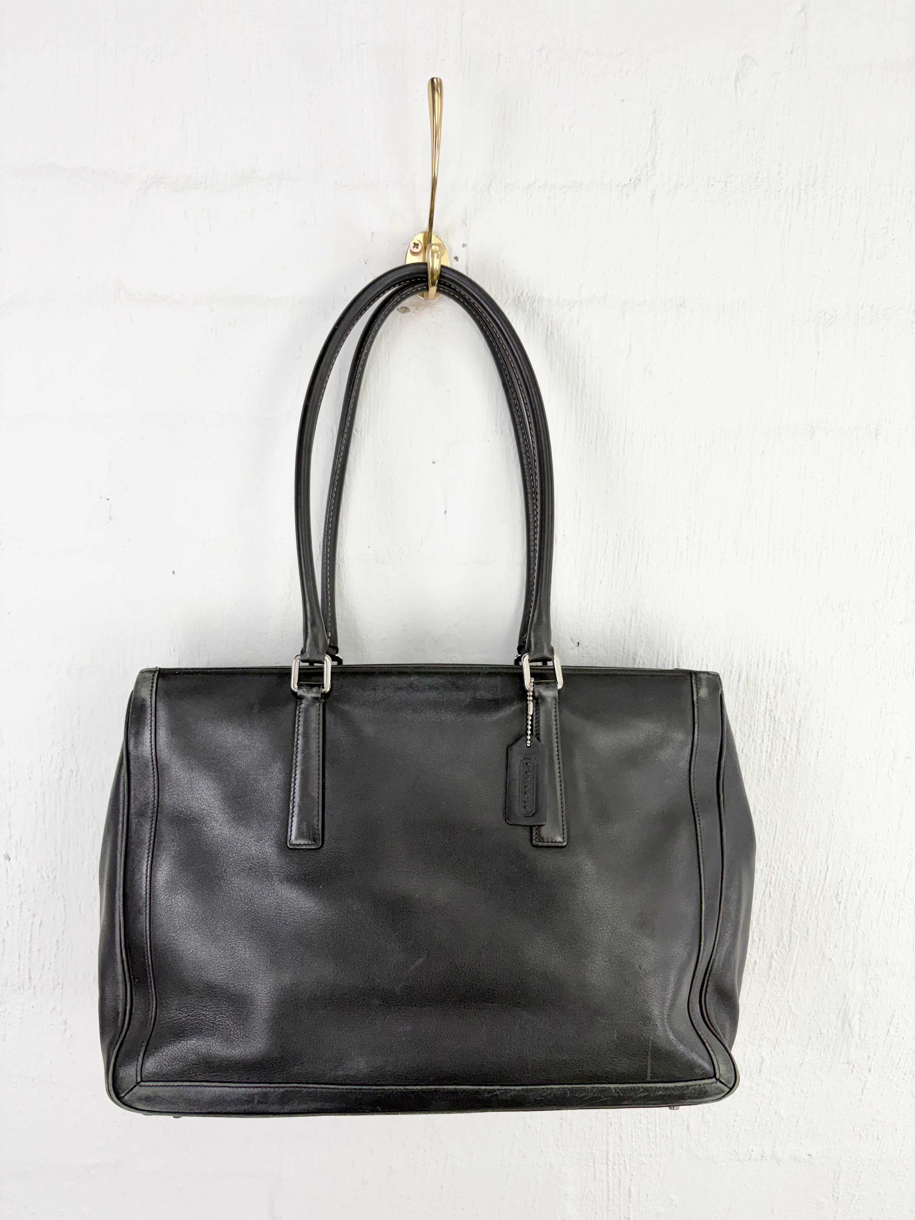 A black leather handbag with double top handles and silver-tone hardware, hanging from a gold hook against a white painted brick wall.