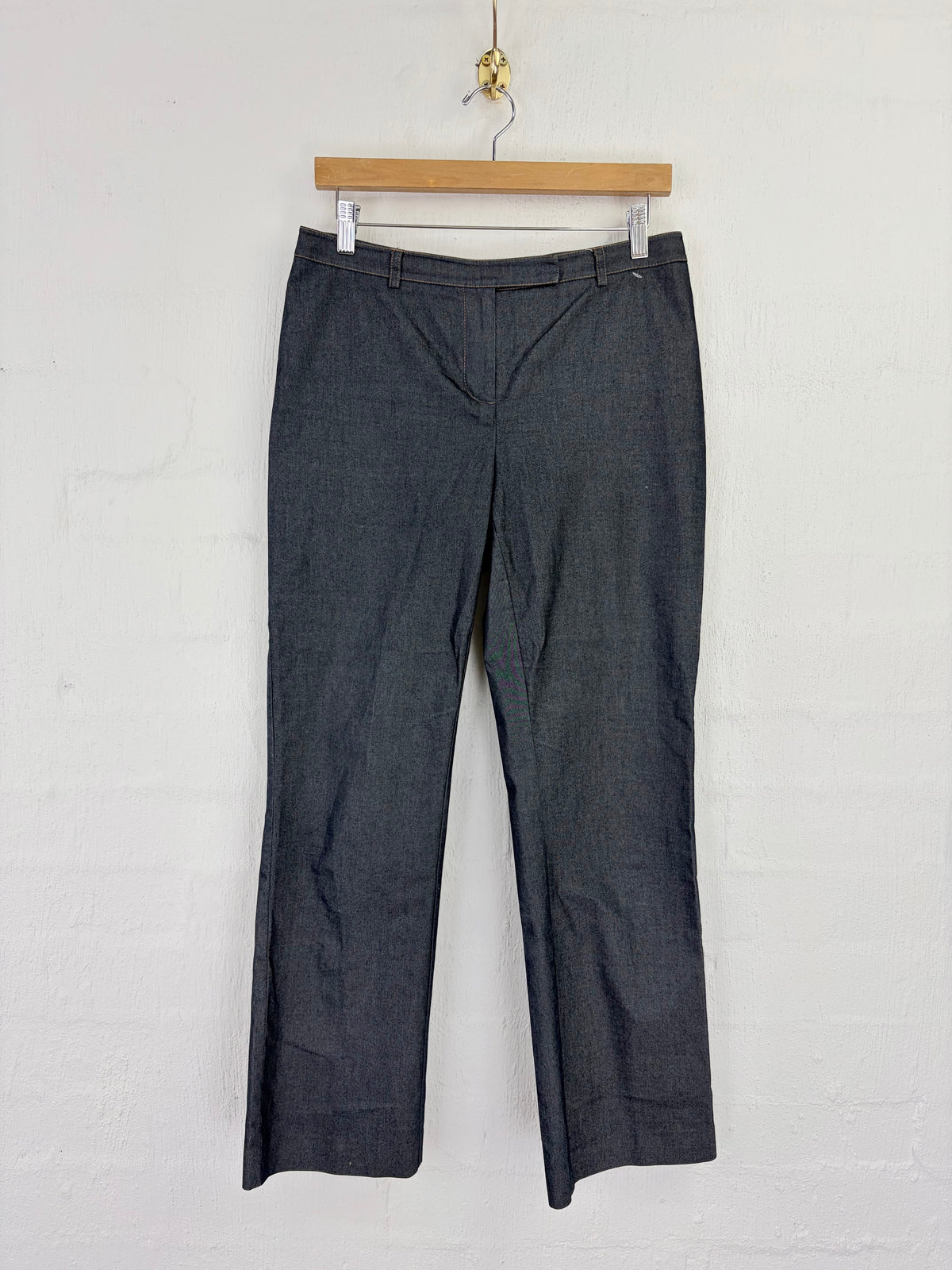 A pair of dark gray cropped trousers hanging from a wooden hanger against a white painted brick wall.
