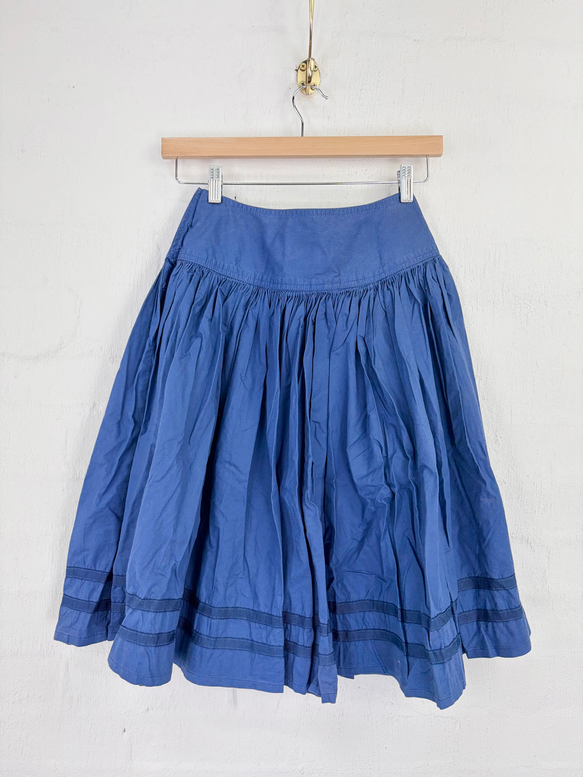 A blue pleated skirt with horizontal dark blue stripe details near the hem, hanging from a wooden hanger against a white textured wall.