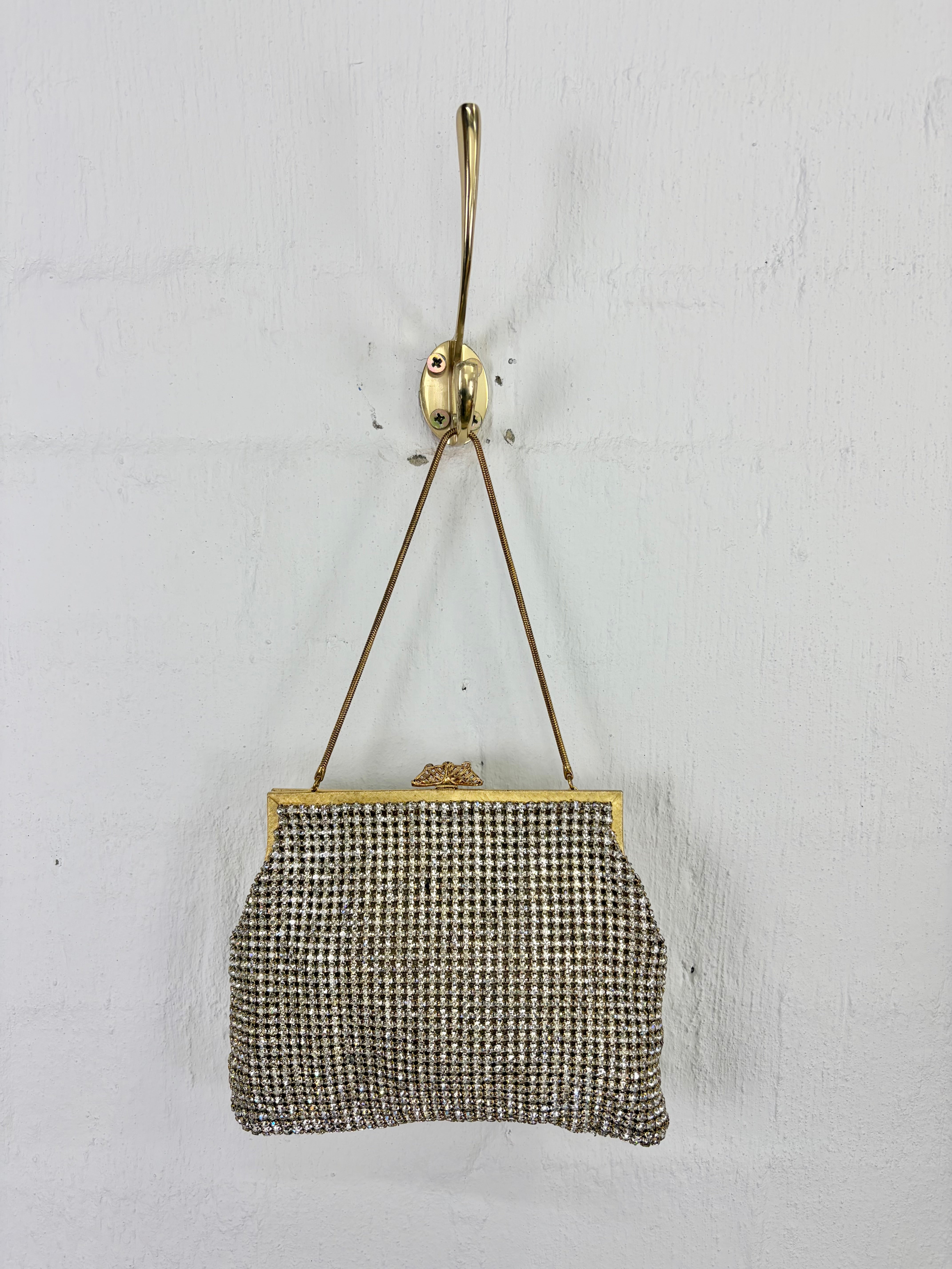A vintage-style clutch purse with a black and beige houndstooth pattern, hanging from a brass hook on a white textured wall.