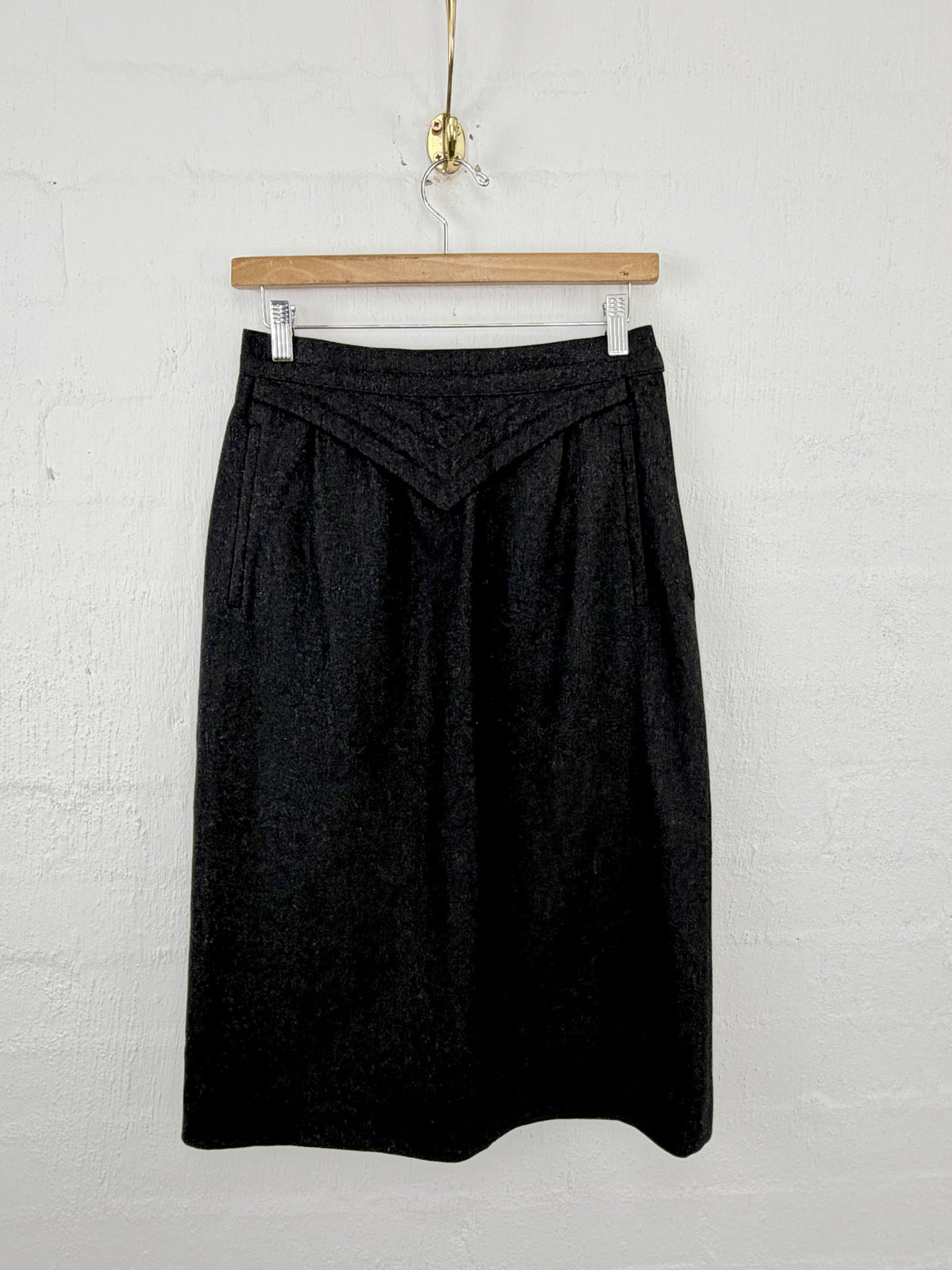 A black textured skirt hanging from a wooden hanger against a white painted brick wall.