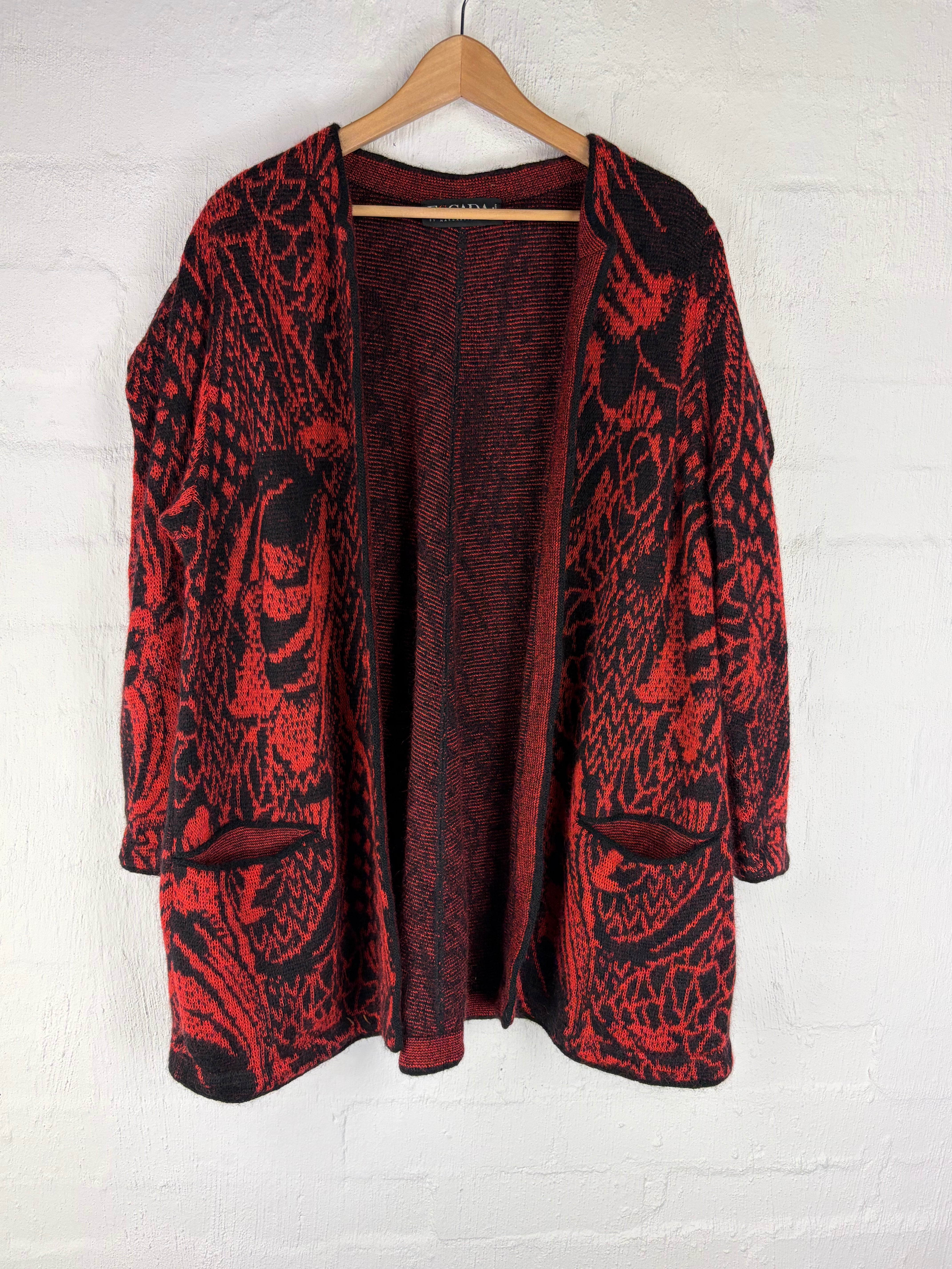 A red and black patterned cardigan sweater with a ribbed knit lining, hanging on a wooden hanger against a white textured wall.