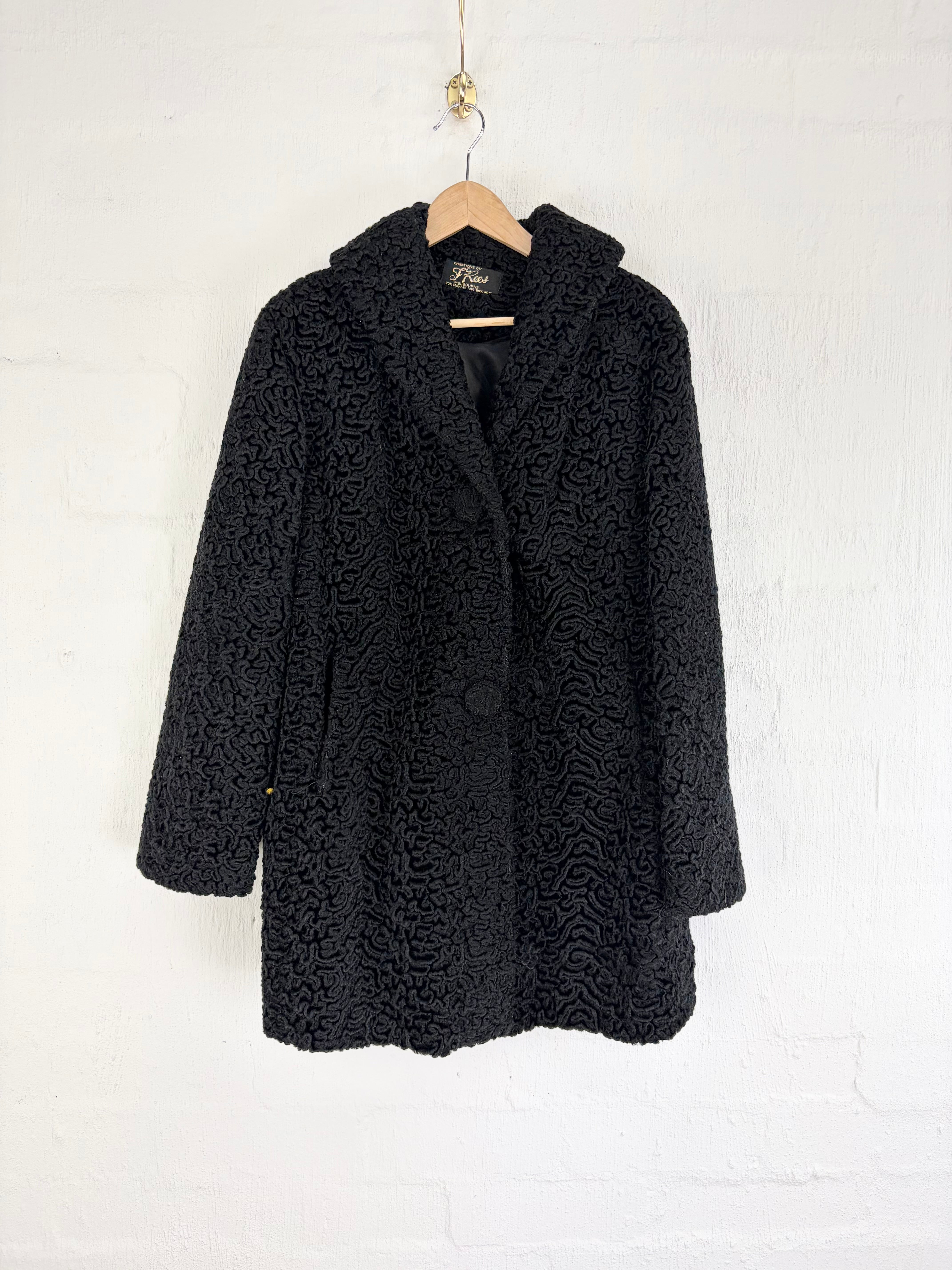 A black textured bouclé coat with a shawl collar, hanging on a wooden hanger against a white painted brick wall.