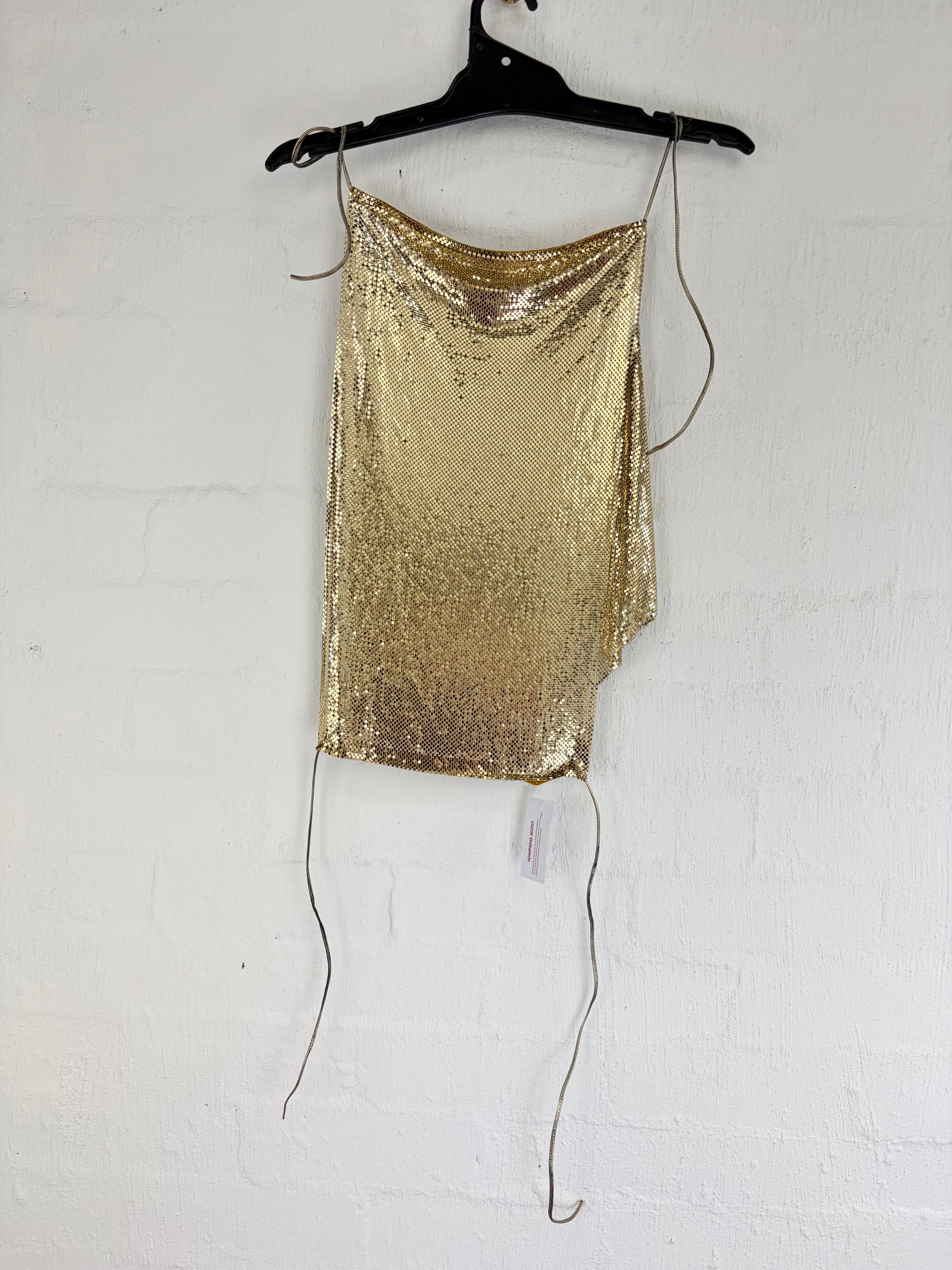 A gold sequined asymmetrical slip dress with thin tie straps, hanging from a black hanger against a white painted brick wall.