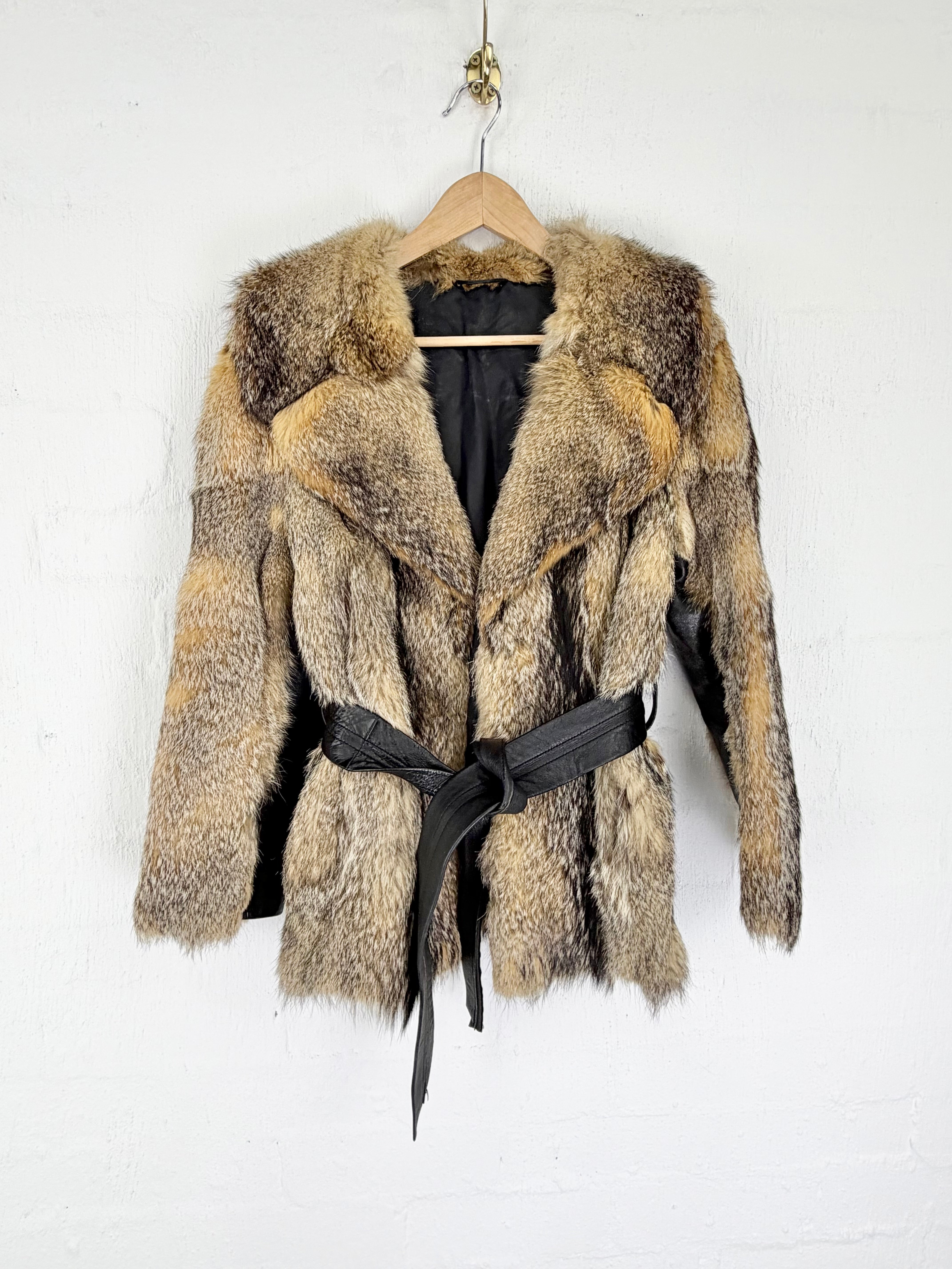 A patchwork fur coat with a black belt, hanging on a wooden hanger against a white painted brick wall.